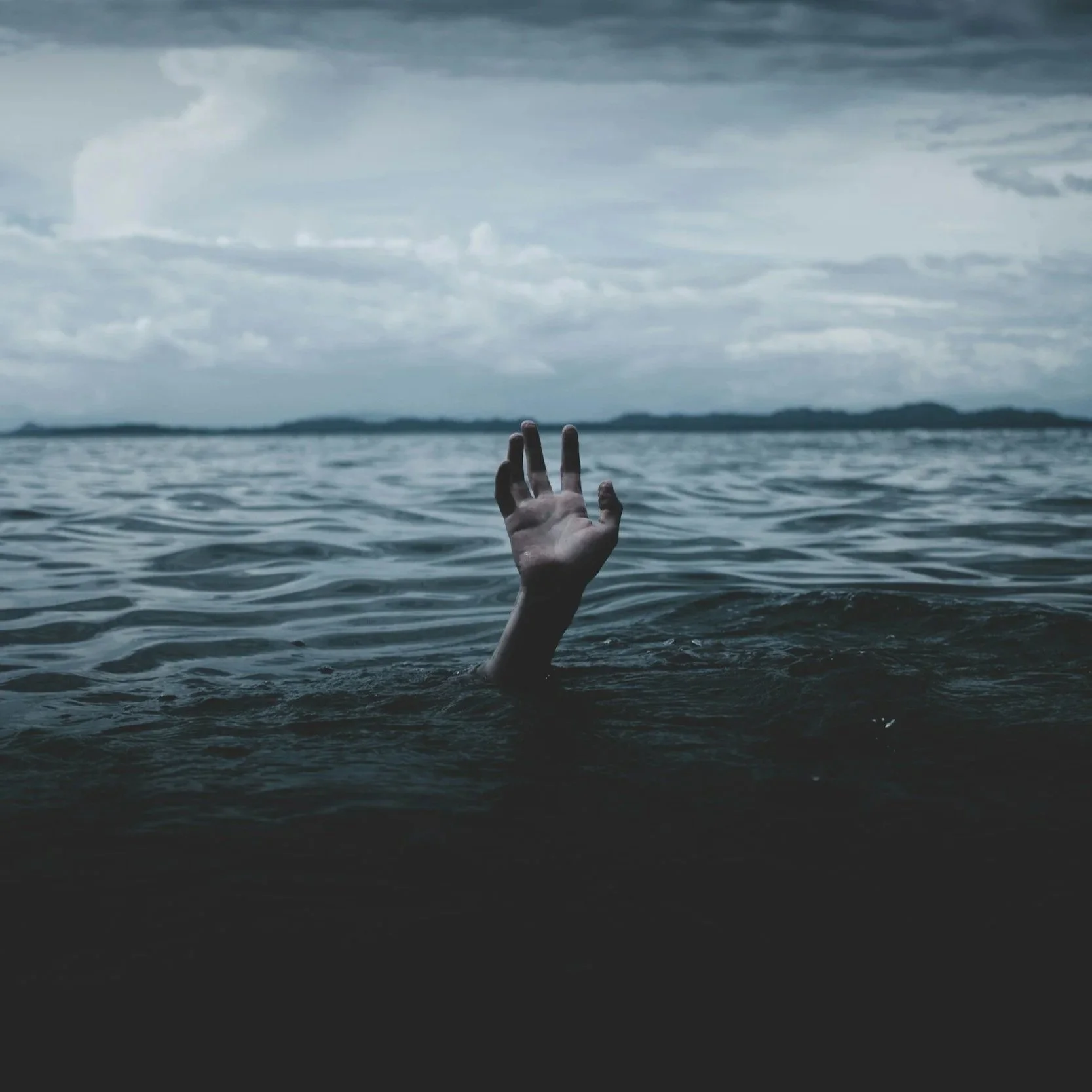 A human hand reaching out of dark water in a lake or ocean during cloudy weather, with distant land visible on the horizon.
