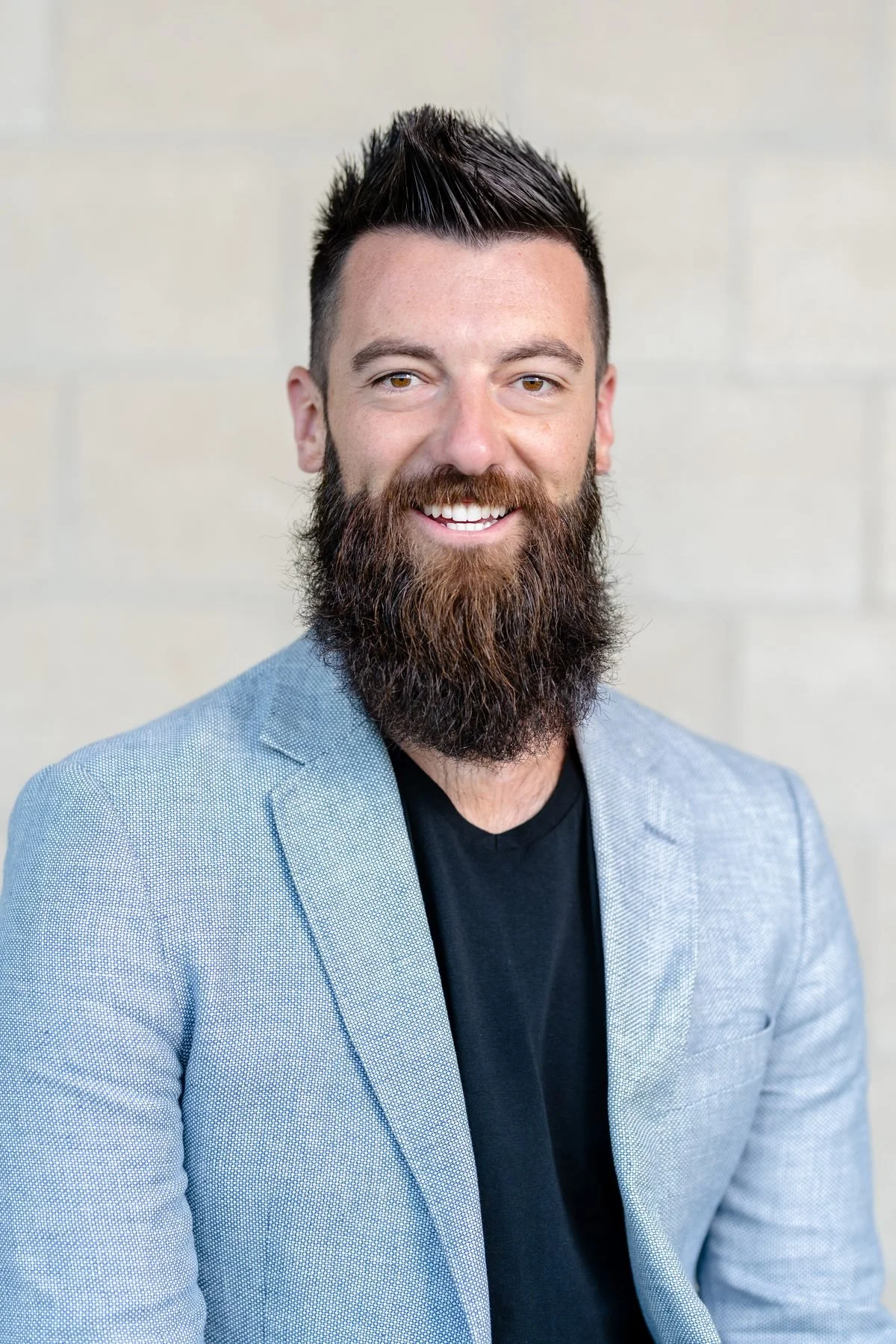Portrait of a smiling man with a beard and short dark hair, wearing a light blue blazer over a black shirt, standing against a neutral background.