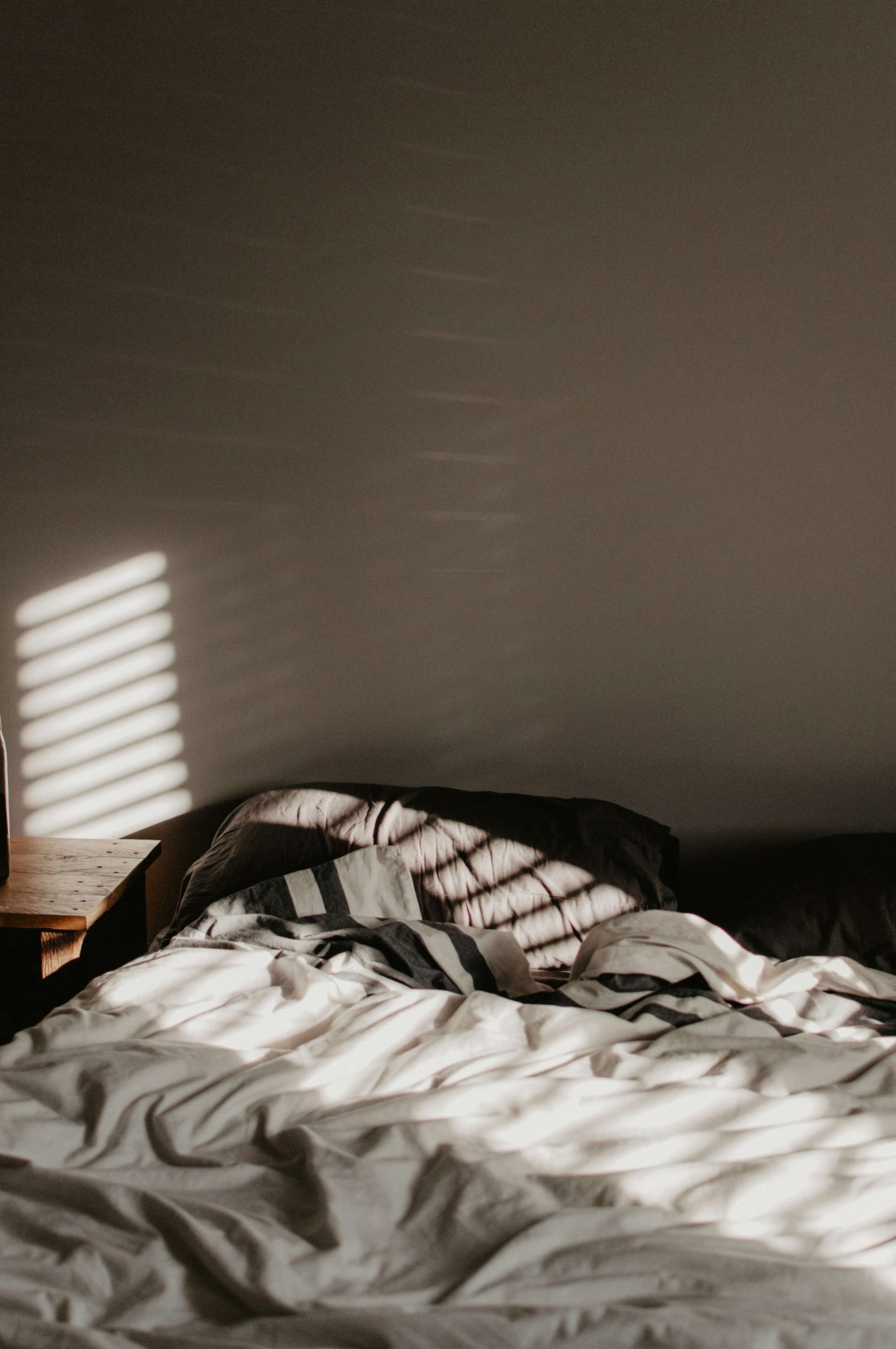 A messy bed with white and patterned sheets, a pillow, and a wooden nightstand in a room with a plain wall and sunlight streaming through blinds.
