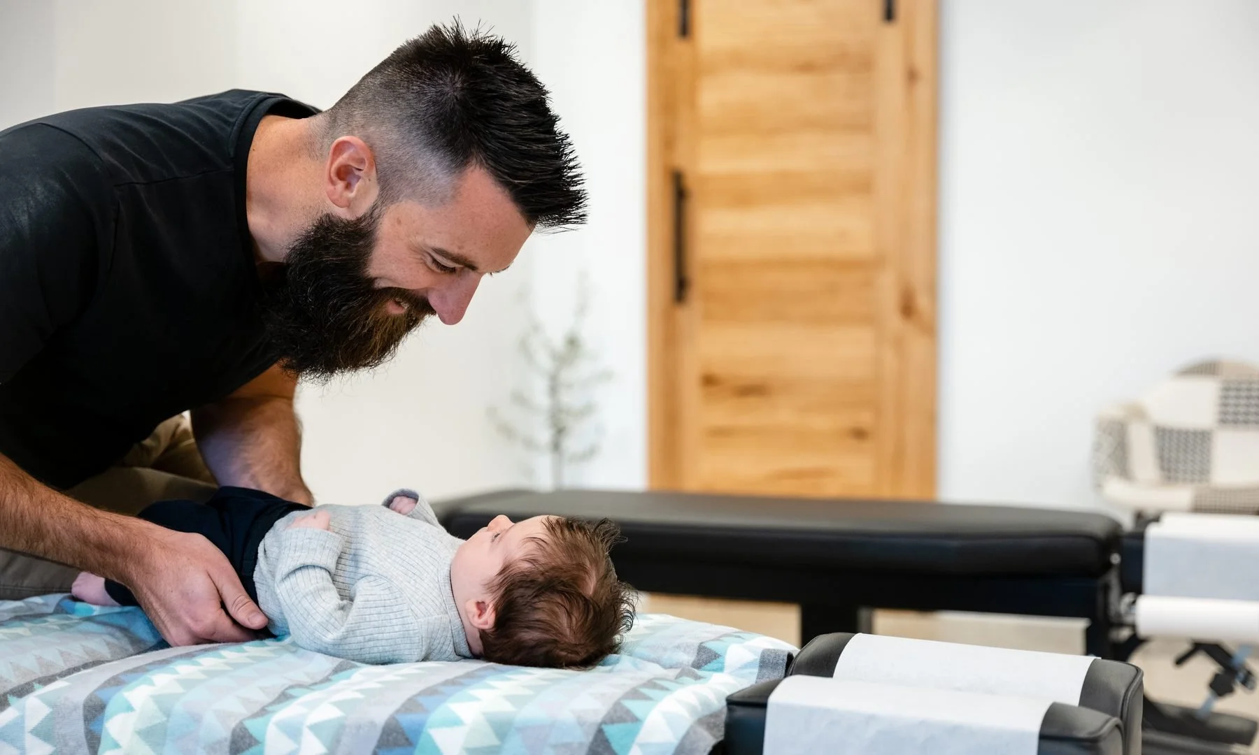 Man with a beard smiling and leaning over a baby lying on a changing table in a room with wooden furniture.