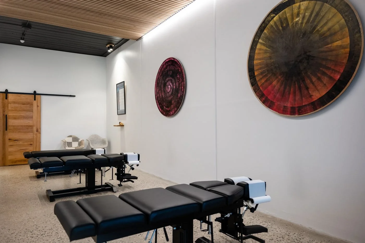 Interior of a wellness or massage therapy room featuring two black adjustable massage tables with face cradles, a wooden sliding barn door, two chairs against a white wall, and abstract circular wall art in red and orange hues.