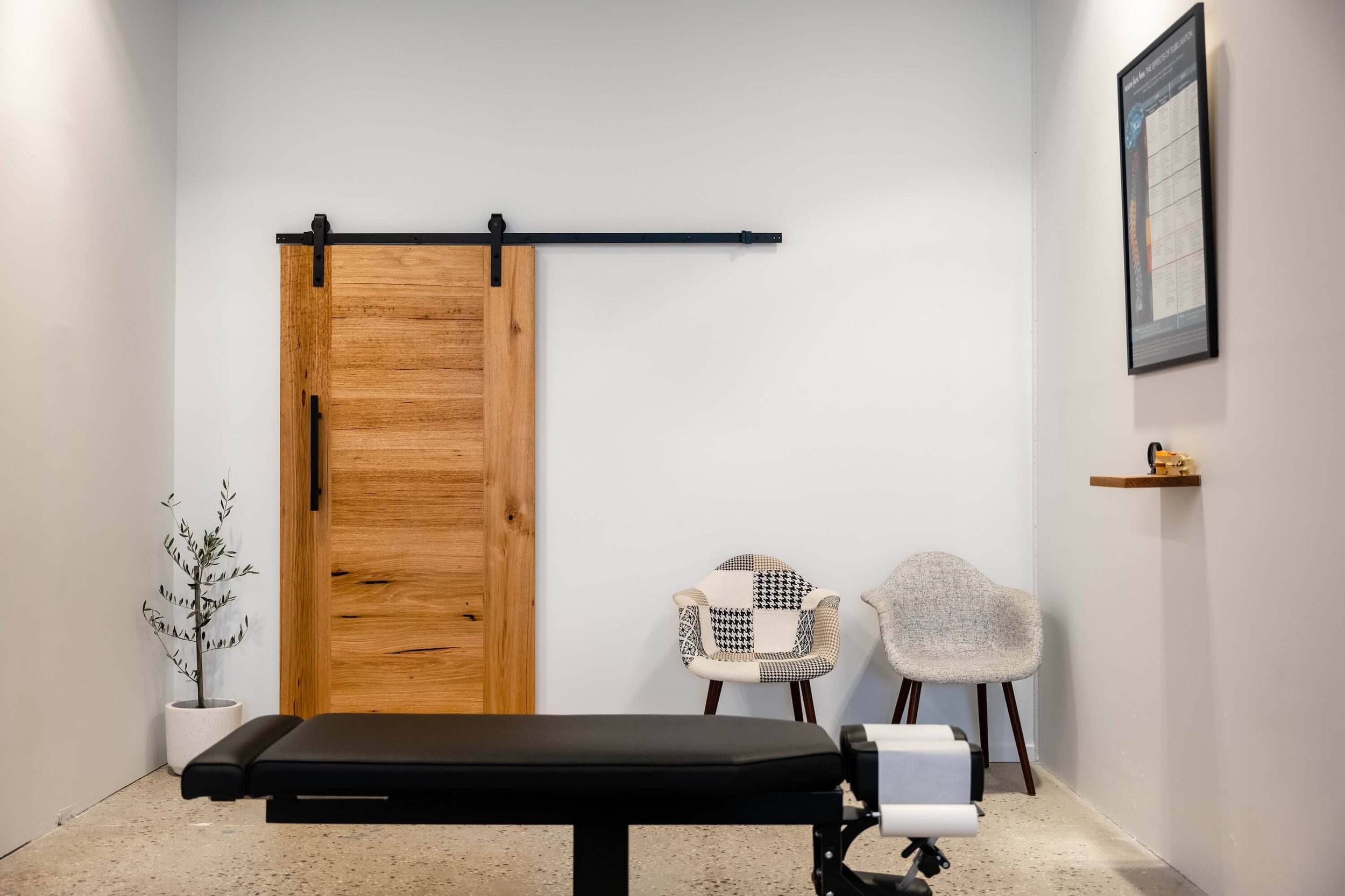 A room with a massage table in the foreground, two chairs with patterned fabric and wood legs, a small floating shelf with a yellow tape measure, a framed chart or poster on the wall, a tall potted plant on the left, and a wooden sliding barn door on the wall.