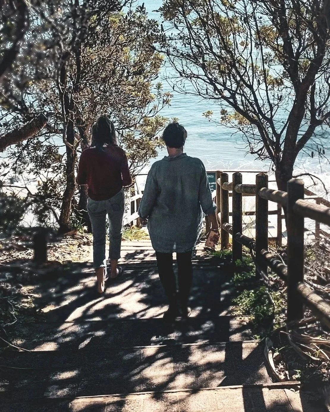 Two people walking down a shaded pathway towards the beach, framed by trees, with water and sandy shore visible in the background.
