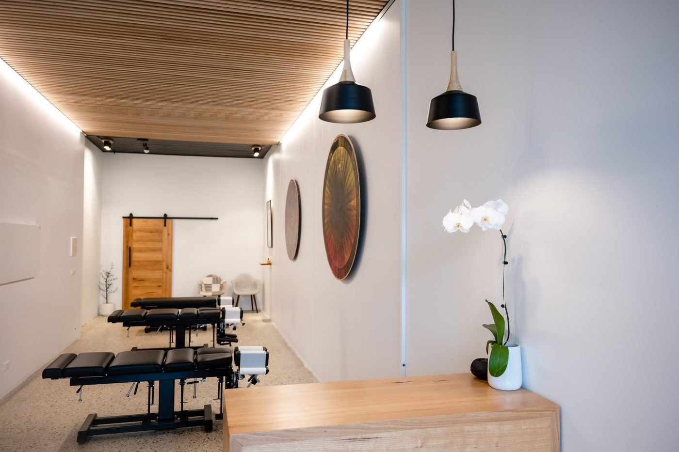 Interior view of a chiropractic or massage therapy clinic with treatment tables, wall art, pendant lighting, and a flower in a white vase.