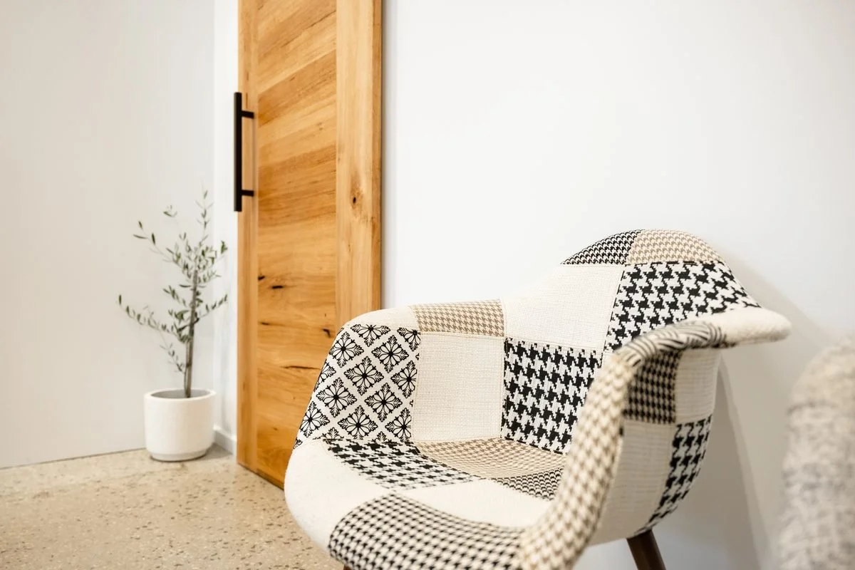 A modern room features a white wall, a wooden sliding door with black handle, a patterned armchair with black and white designs, and a potted plant near the corner.