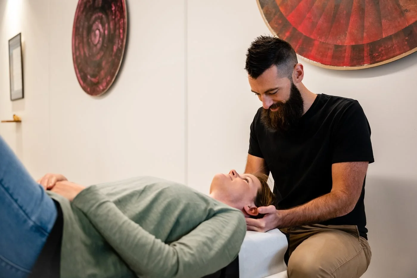 A man providing cranial therapy to a woman lying on a therapy table in a room with abstract wall art.
