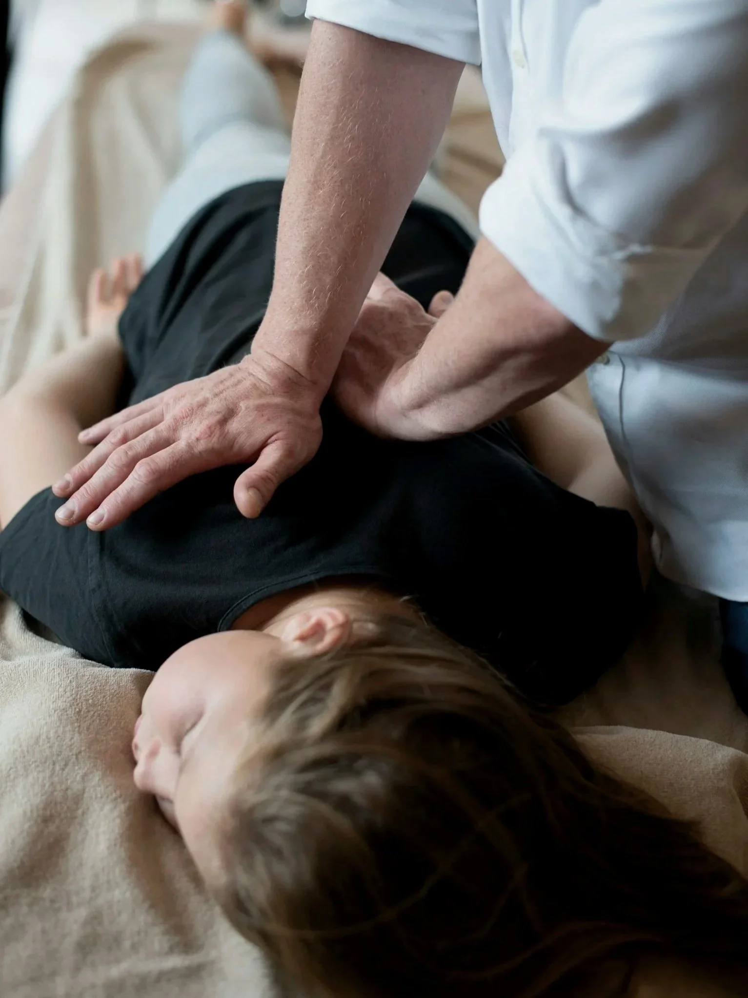 A person performing CPR or chest compressions on a child lying on a soft surface, with the child's face turned to the side and eyes closed.