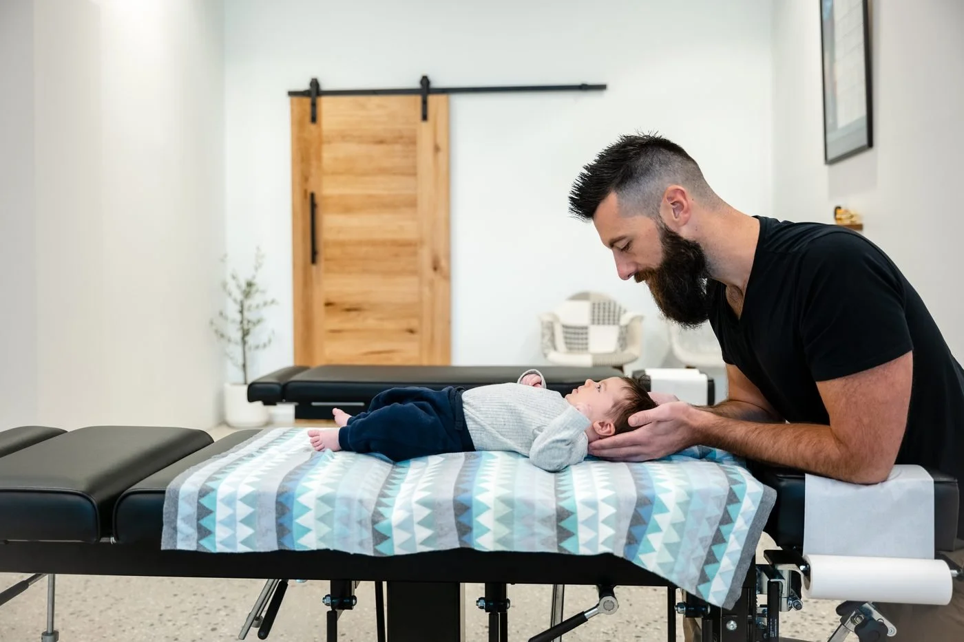 A man with a beard and black shirt looking at a baby lying on a medical examination table in a bright room.