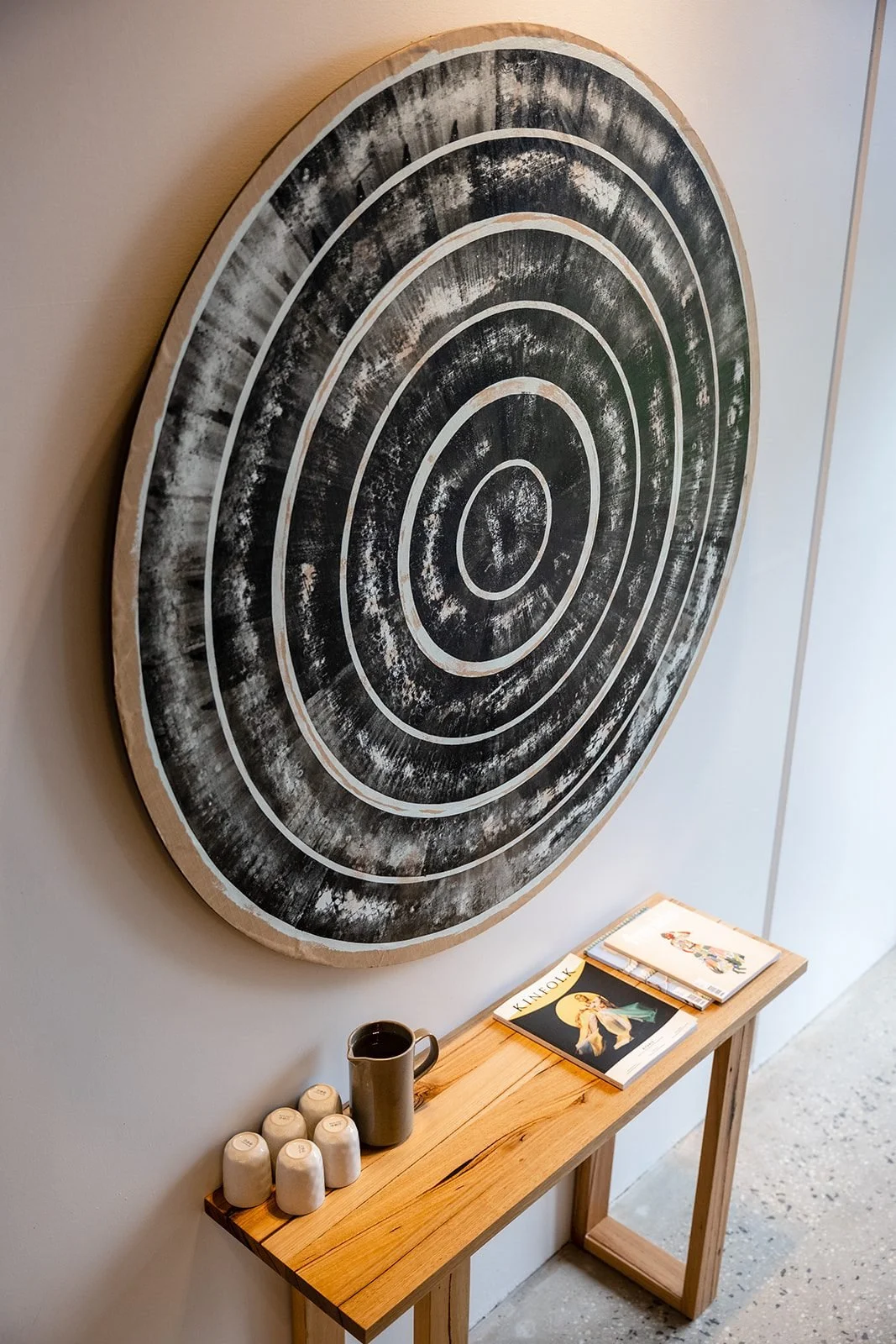 A wooden console table with ceramic cups, a mug, and magazines; large round wall art with concentric circles in black and white.