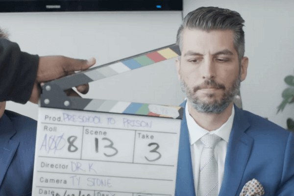 A man in a blue suit and white shirt looks serious as a clapperboard is held in front of him on a film set.