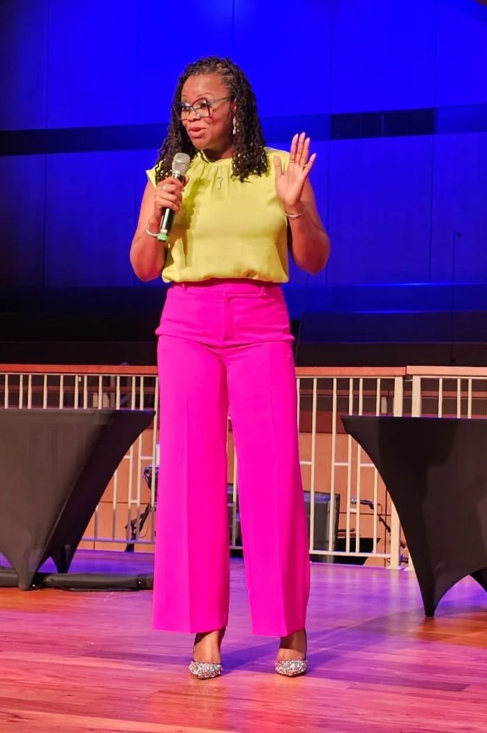 A woman speaking on stage, holding a microphone, wearing a yellow top, bright pink pants, and silver glitter heels, with her right hand raised in a gesture.