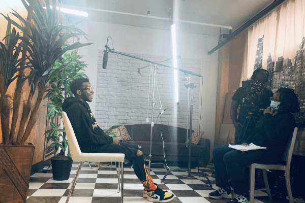 A man in a recording studio being interviewed by two women, one holding a notebook and pen, while the other works with audio equipment. The studio has a checkered floor, a potted plant, a black couch, and a cityscape mural on the wall.