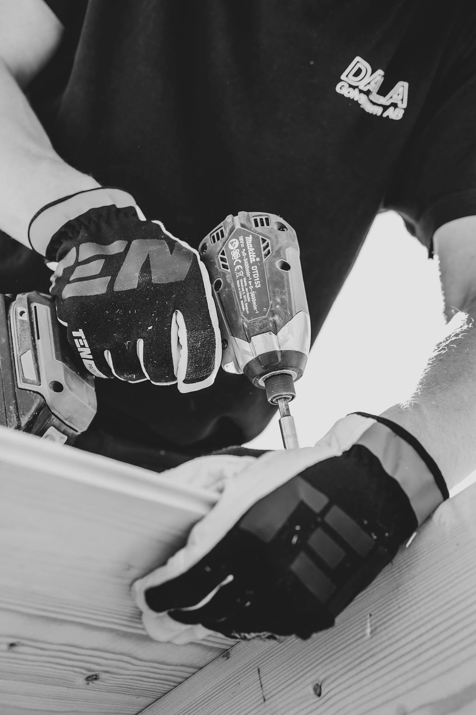 A person wearing gloves and a black shirt with the logo 'DALA Communication AB' is using a cordless drill to work on a wooden surface.