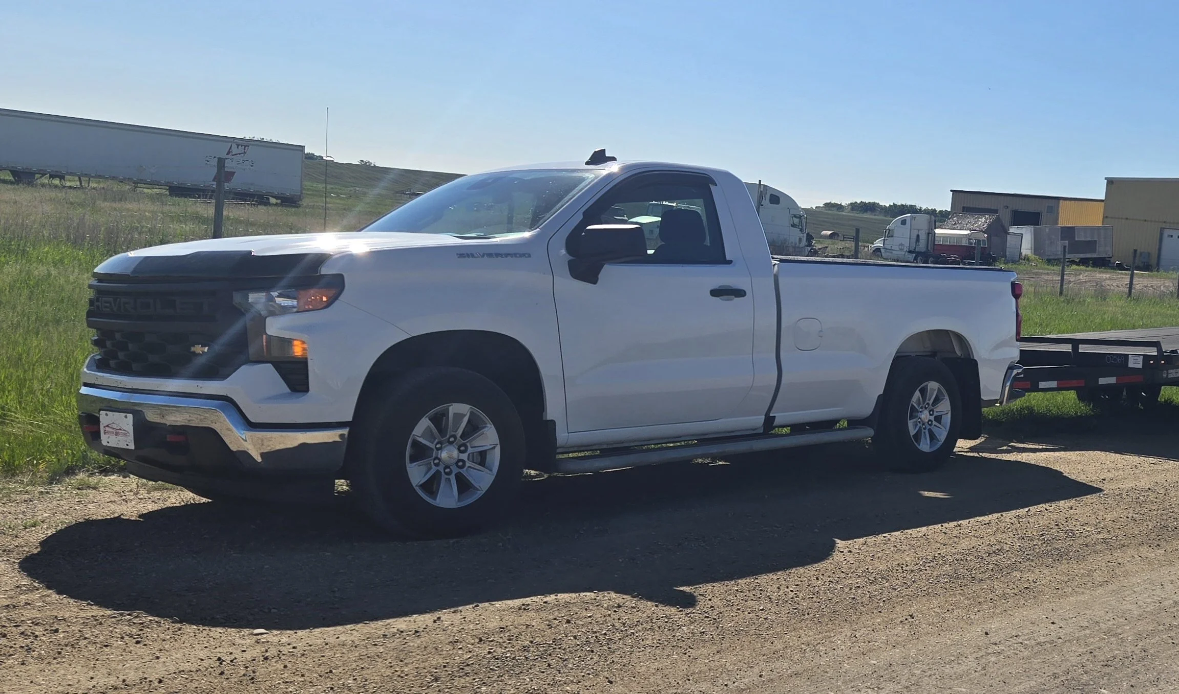 White Chevrolet Silverado pickup truck parked on a gravel surface near grassy field, with farm buildings and semi-trailers in the background under a clear blue sky.