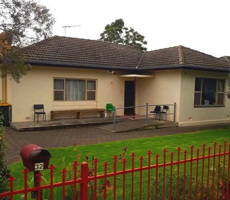 A single-story house with a brown tiled roof and beige exterior walls. There is a small patio with a bench, two black chairs, and a green chair. The house has windows with security screens. A red fence surrounds a lawn in front, and a red mailbox with the number 32 is nearby.