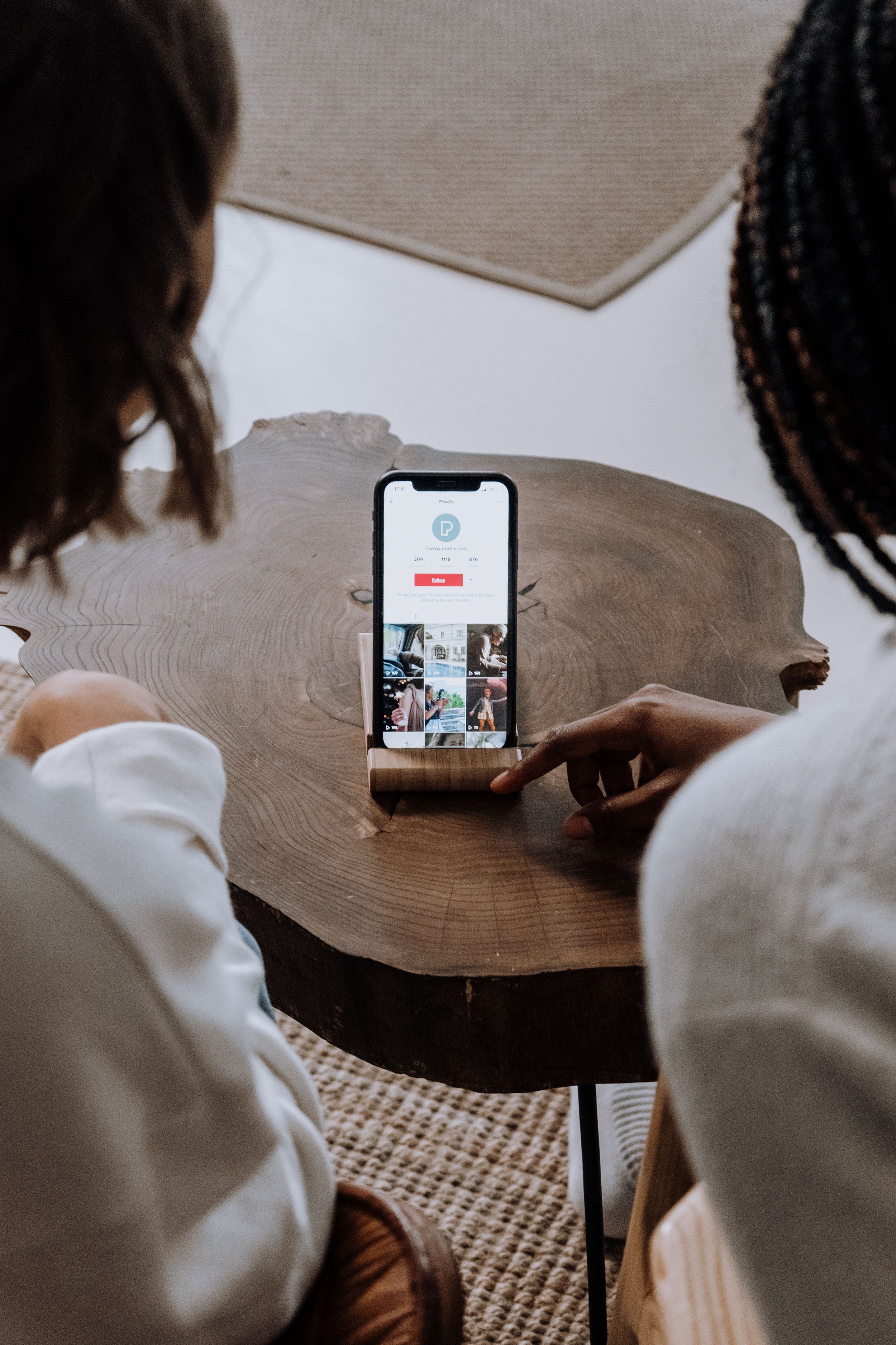 Two people are sitting at a round wooden table looking at a smartphone, which displays a photo application, with their focus on the phone.