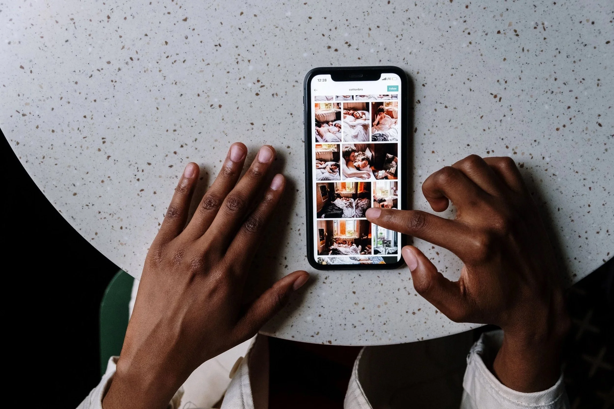 A person is holding a smartphone displaying a grid of bed photos in the Instagram app on a speckled white table.