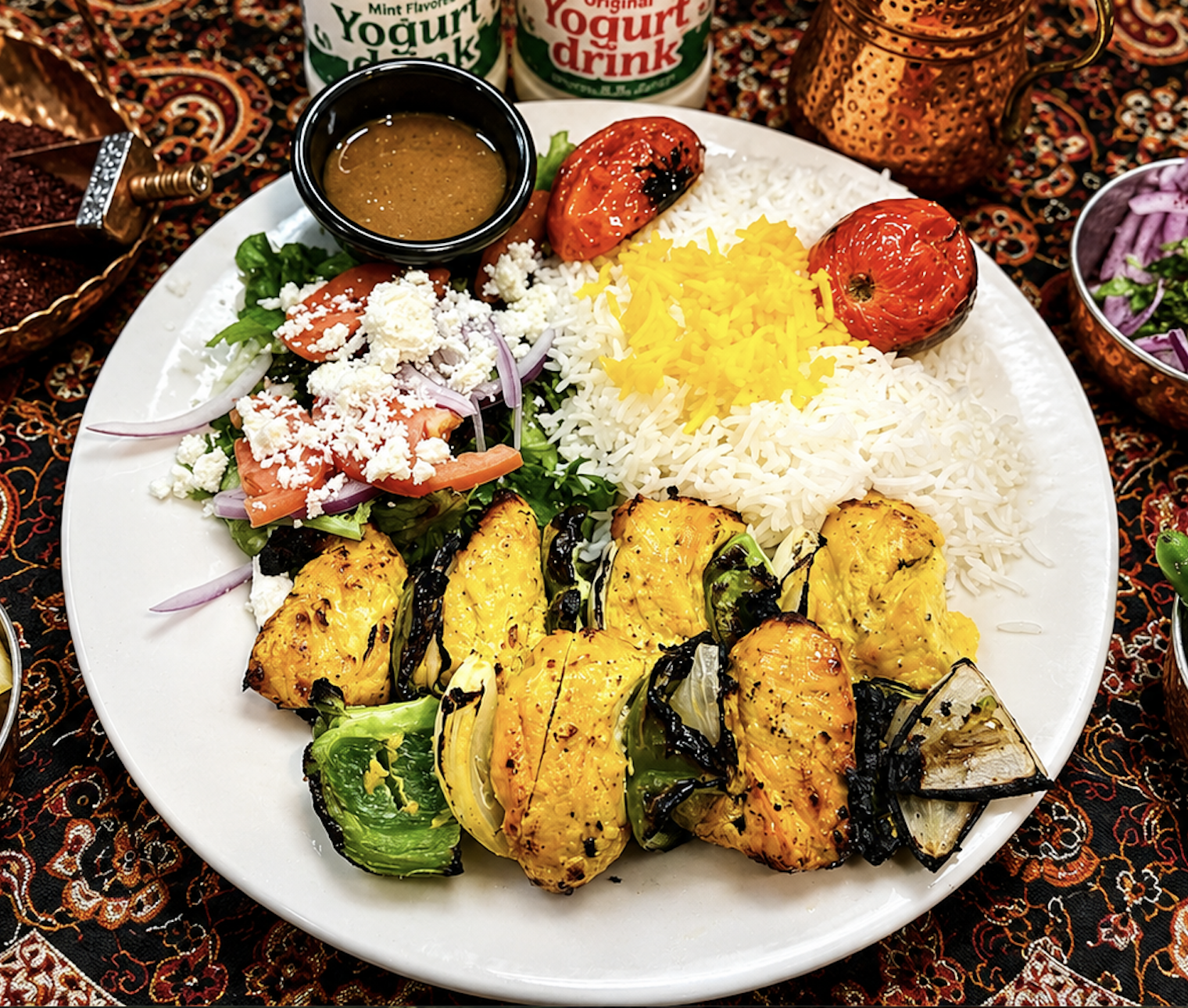 A large white plate contains grilled chicken with a yellow marinade, steamed white rice with shredded yellow cheese, a fresh salad with lettuce, tomato, onion, and feta cheese, and grilled tomatoes.