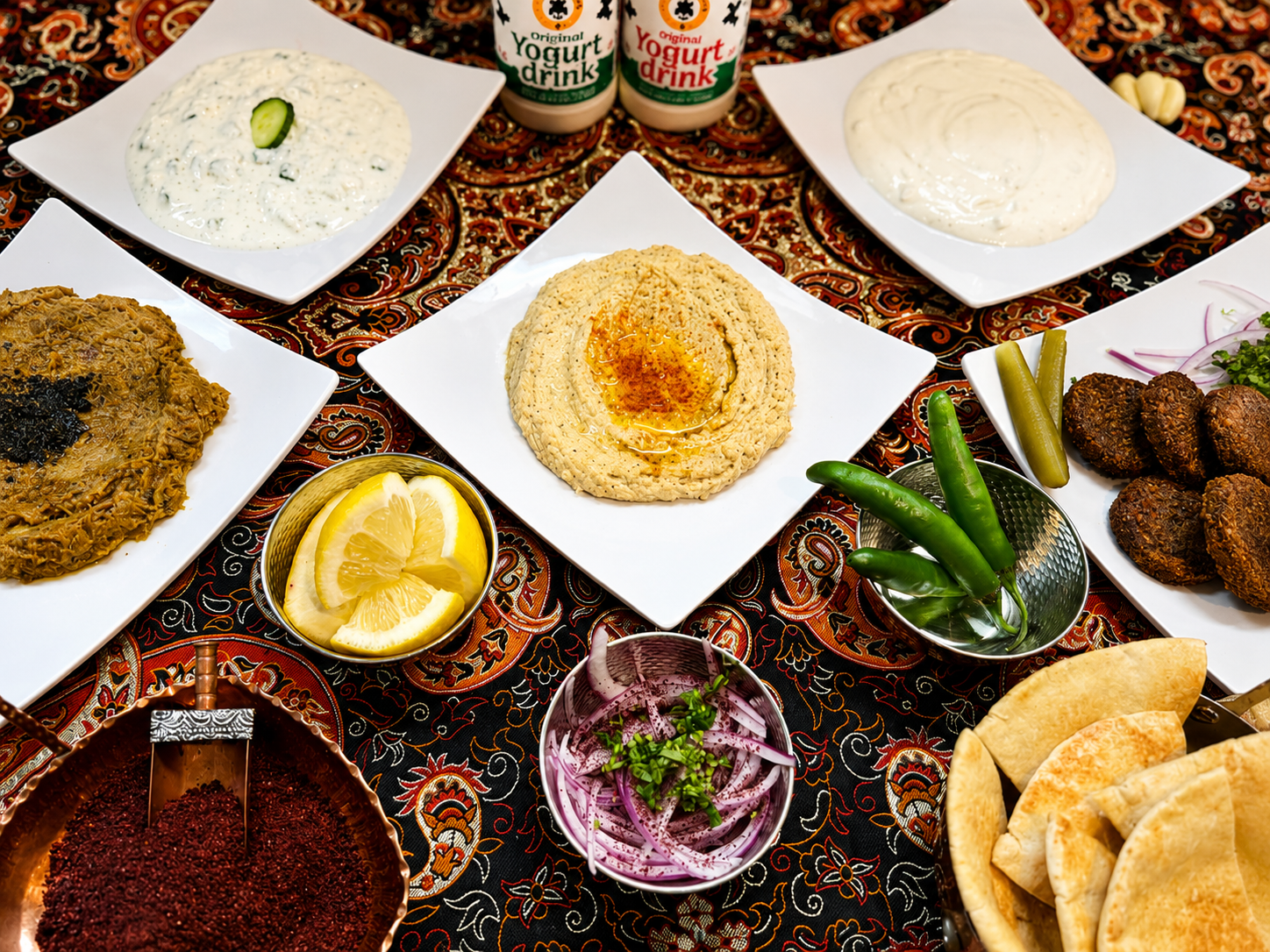 A colorful Middle Eastern meal spread on a table with various dishes including hummus, falafel, tabbouleh, pickles, lemon slices, yogurt, and pita bread, along with hot sauce and condiments.