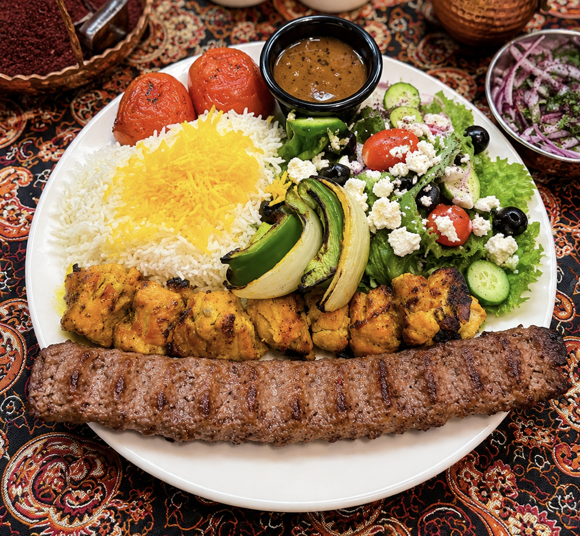 Plate of Middle Eastern food with rice, grilled meat, grilled vegetables, salad, tomatoes, stuffed grape leaves, and a small bowl of sauce, on a patterned tablecloth.