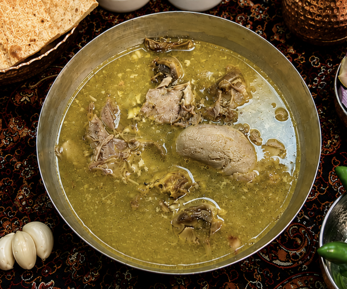A metal bowl filled with traditional chicken soup containing chicken organs and broth, surrounded by garlic, bread, and condiments on a decorative tablecloth.