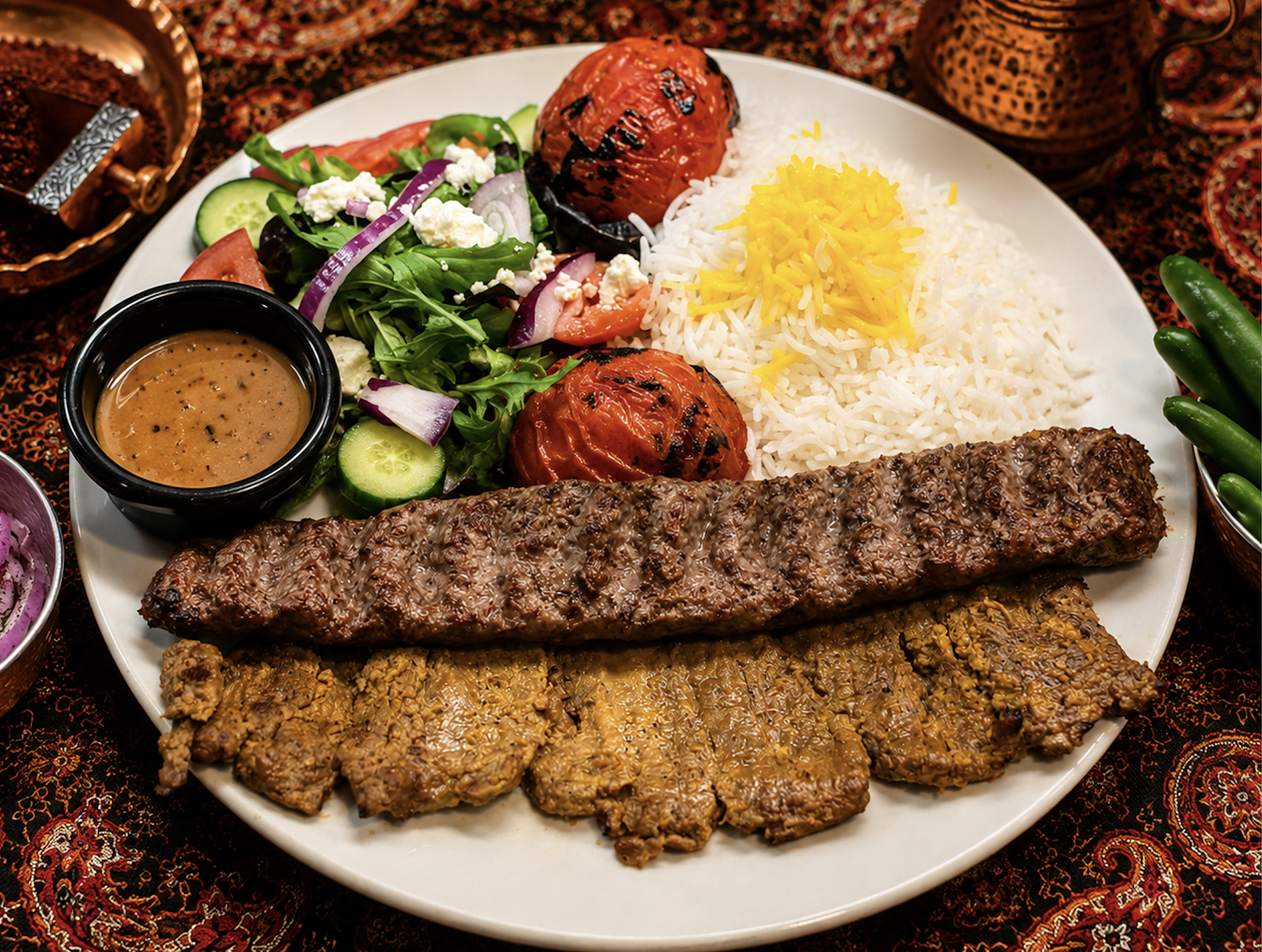 A plate of Middle Eastern food with rice, grilled meat, tomato, cucumber, salad, stuffed grape leaves, and saffron rice.