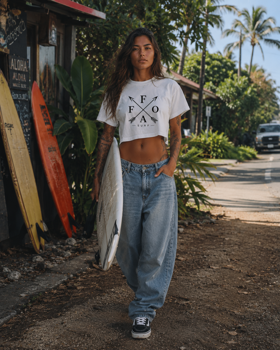 A woman with tattoos and long hair walking along a tropical street while holding a surfboard. She is wearing a crop top and baggy jeans.