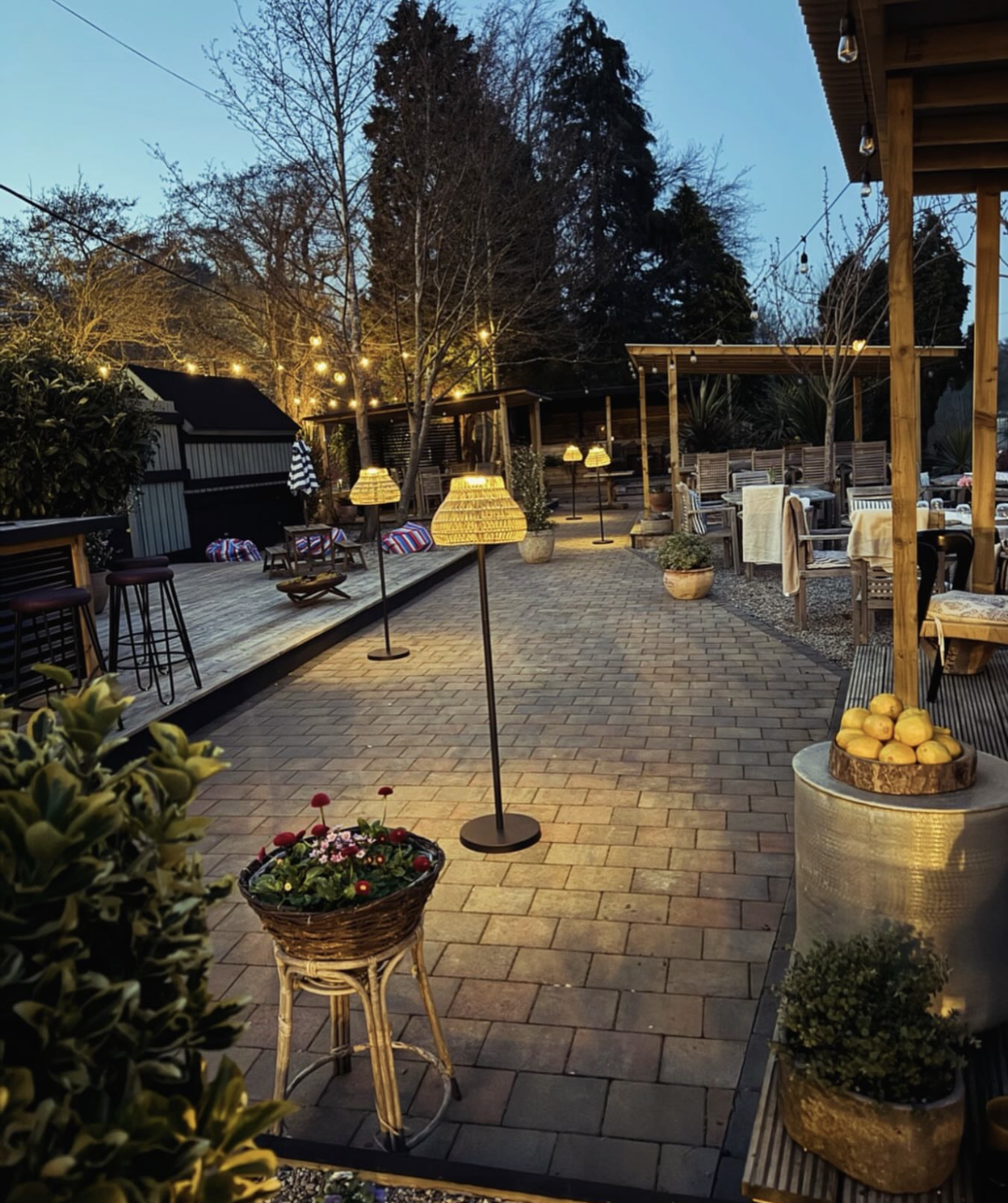 Outdoor patio area with string lights, potted plants, wooden furniture, and a paved walkway, during dusk.
