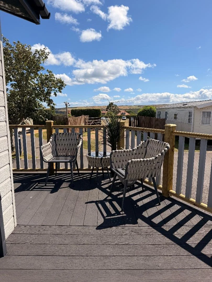 A wooden deck with three woven chairs and a potted plant under a blue sky with scattered clouds in a suburban backyard.