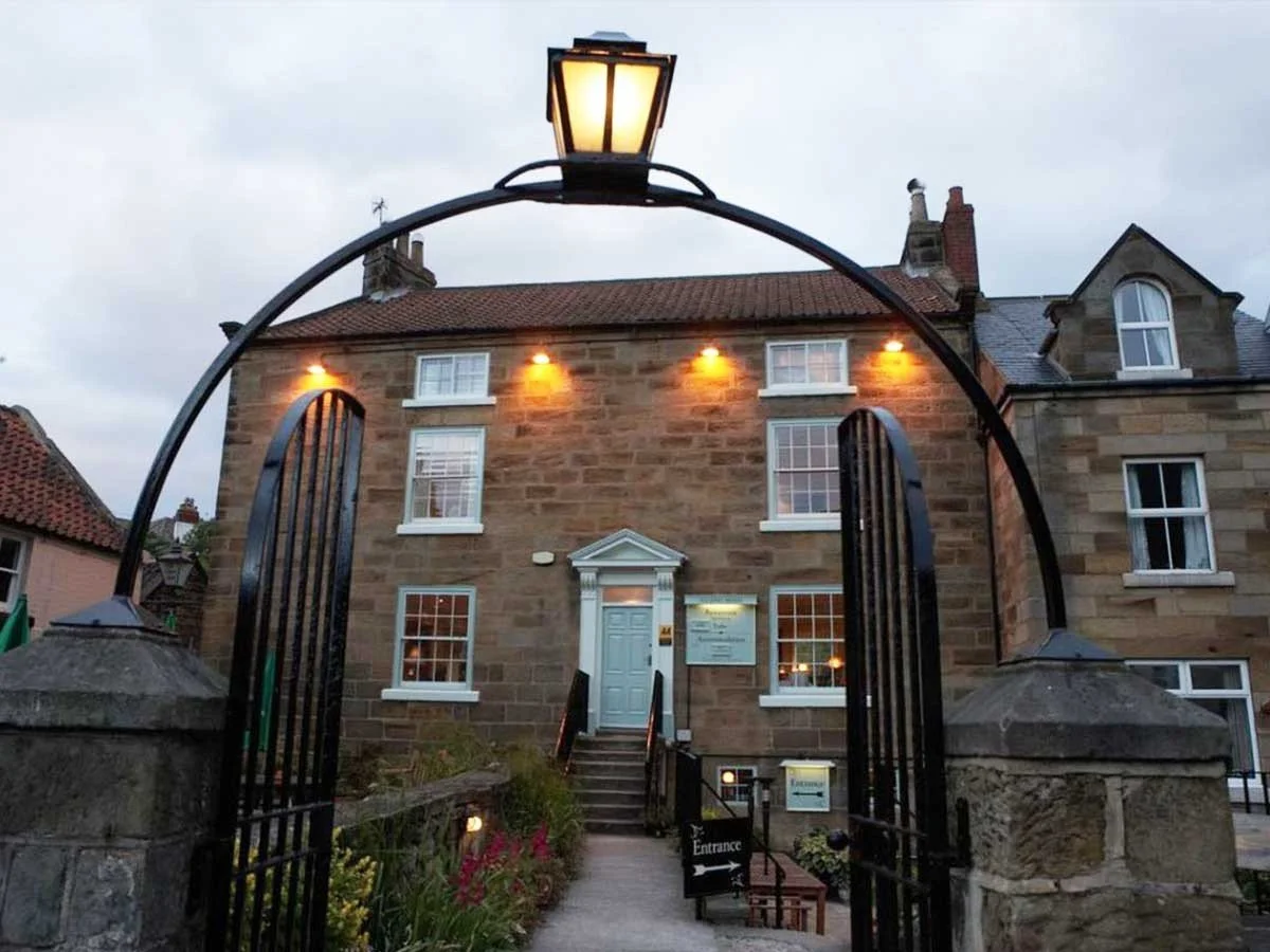 A stone building seen through a black wrought iron gate with a lit street lamp on top. The building has multiple windows with white frames and exterior lights, and a white door with steps leading up to it. There is a small garden with flowers in front.