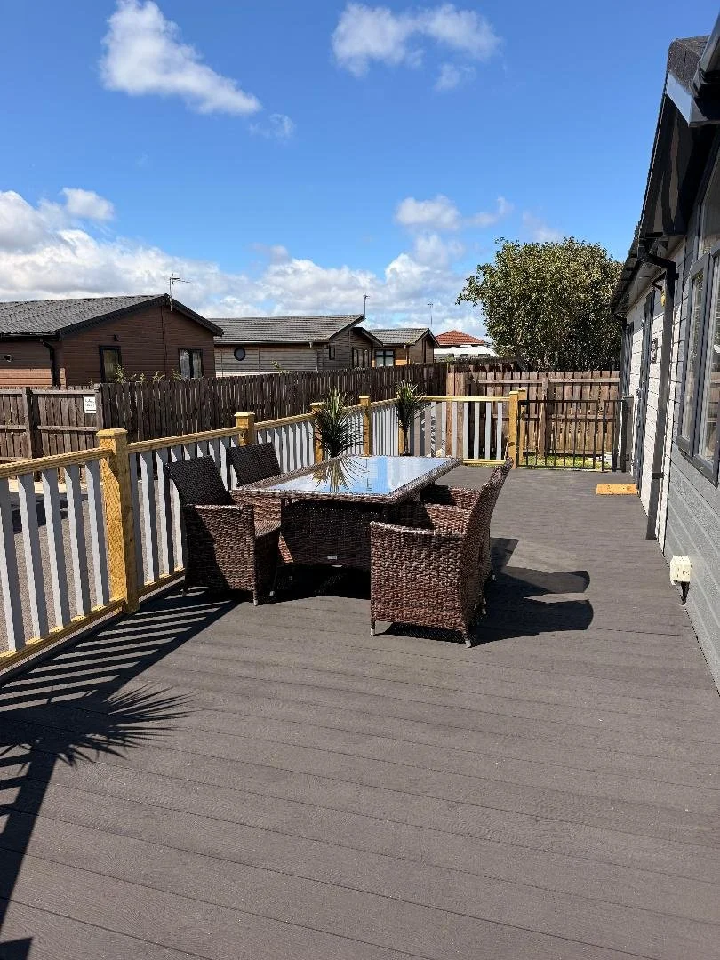 Outdoor deck with wicker table and six wicker chairs, surrounded by a wooden fence, with neighboring houses and a tree in the background, under a partly cloudy sky.