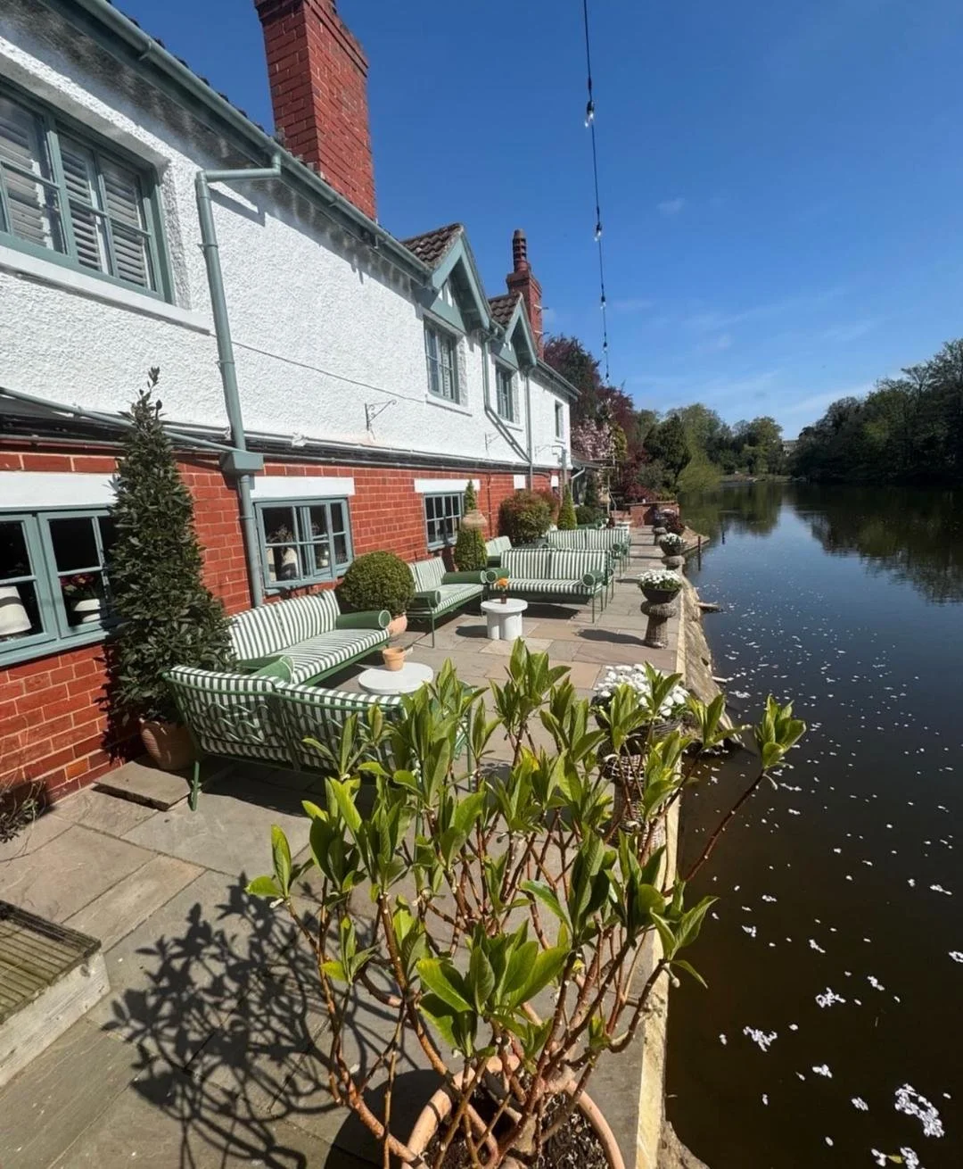 A patio with striped green and white outdoor furniture next to a river, with potted plants and decorative urns along the edge, and a house with white and brick walls and multiple windows in the background under a clear blue sky.