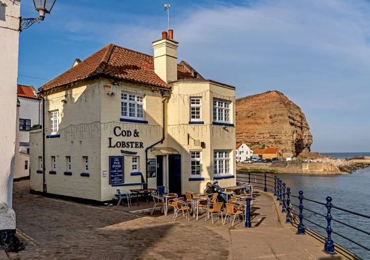 A white building with a red tile roof labeled 'Cod & Lobster,' with outdoor tables and chairs along a waterfront, and a hill with a rocky formation in the background.