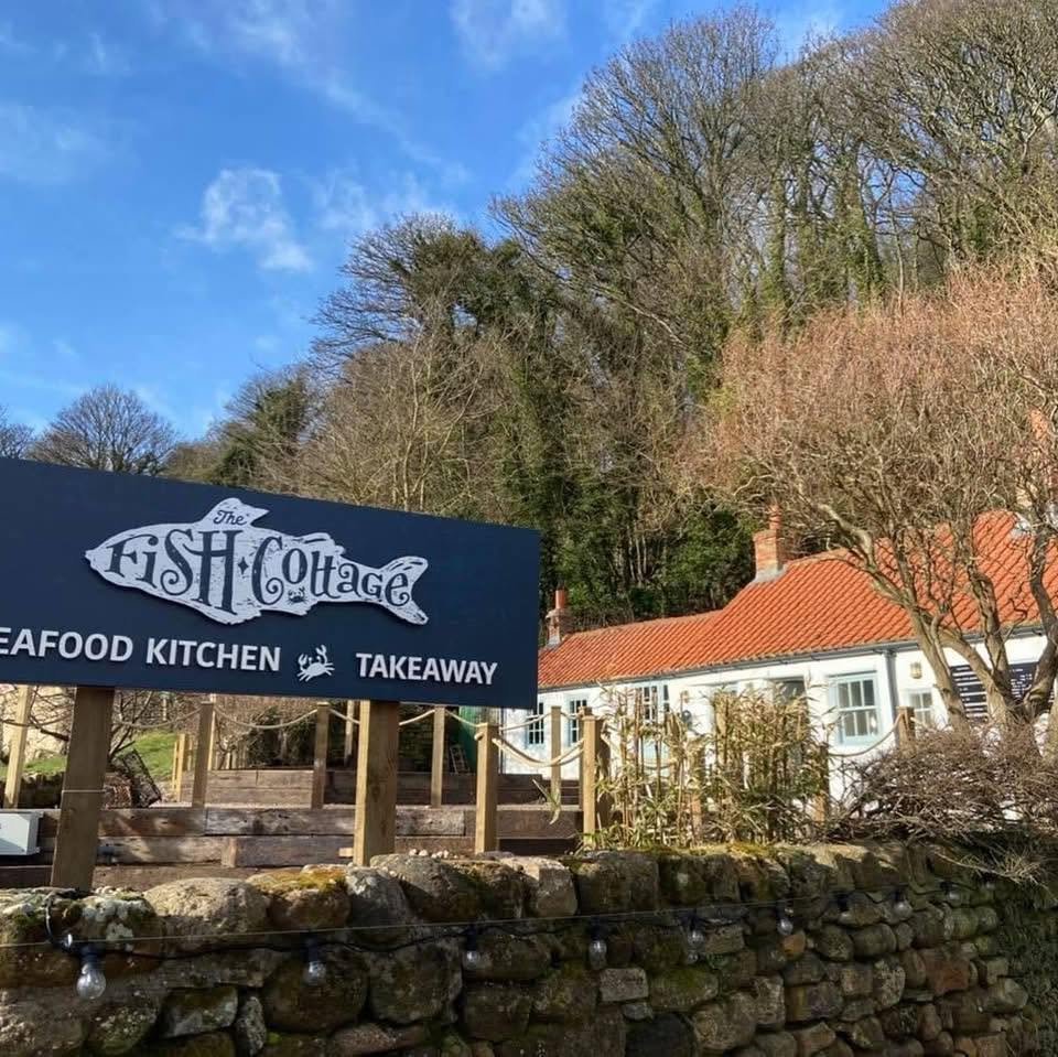 A sign for The Fish Cottage restaurant, which offers seafood cuisine and takeout, with a house and trees in the background under a blue sky.