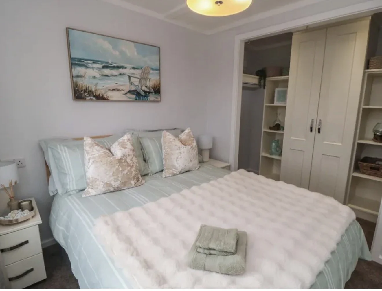 A cozy bedroom with a bed covered in striped bedding and a fluffy white throw blanket. Four pillows, two with a textured pattern, are arranged on the bed. A towel is folded on the blanket. There are two nightstands with lamps and decorative items. A 