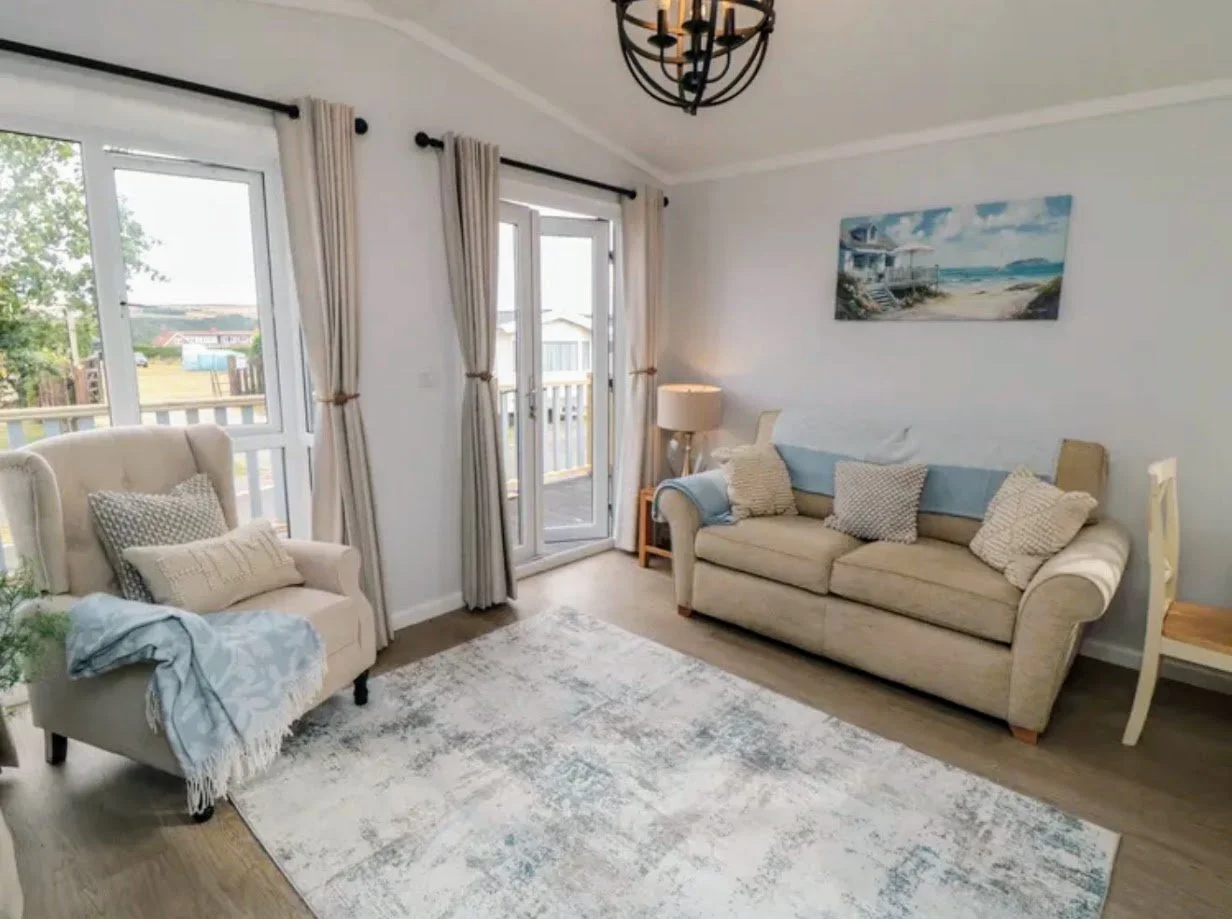 Living room with two beige sofas, a beige armchair, a white rug, a large window with curtains, a sliding glass door, a floor lamp, a ceiling light fixture, and wall art of a beach scene.