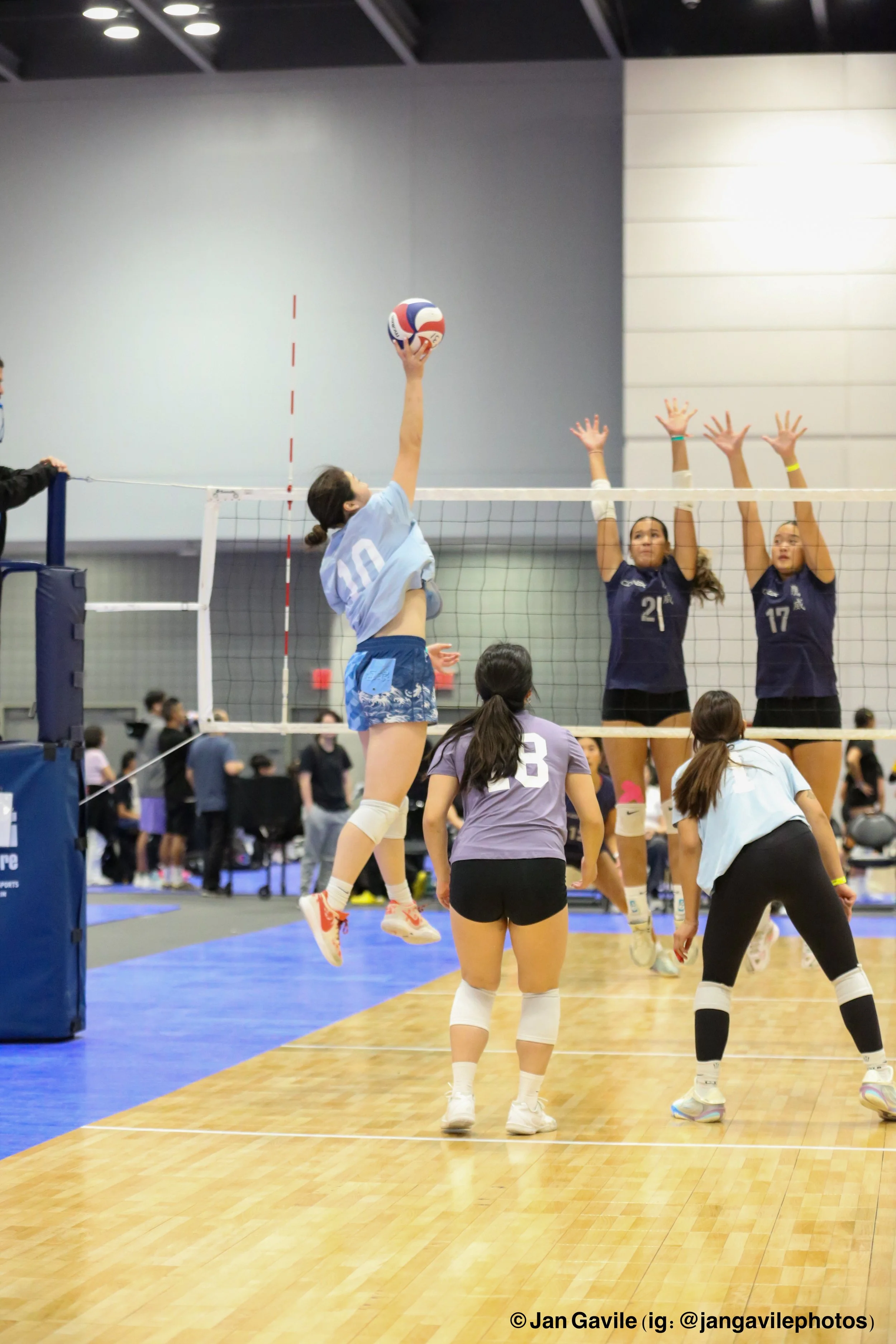 A volleyball game in progress with players at the net, including one player jumping to hit the ball and two players on the opposing side preparing to block. The scene is inside a sports hall with spectators in the background.