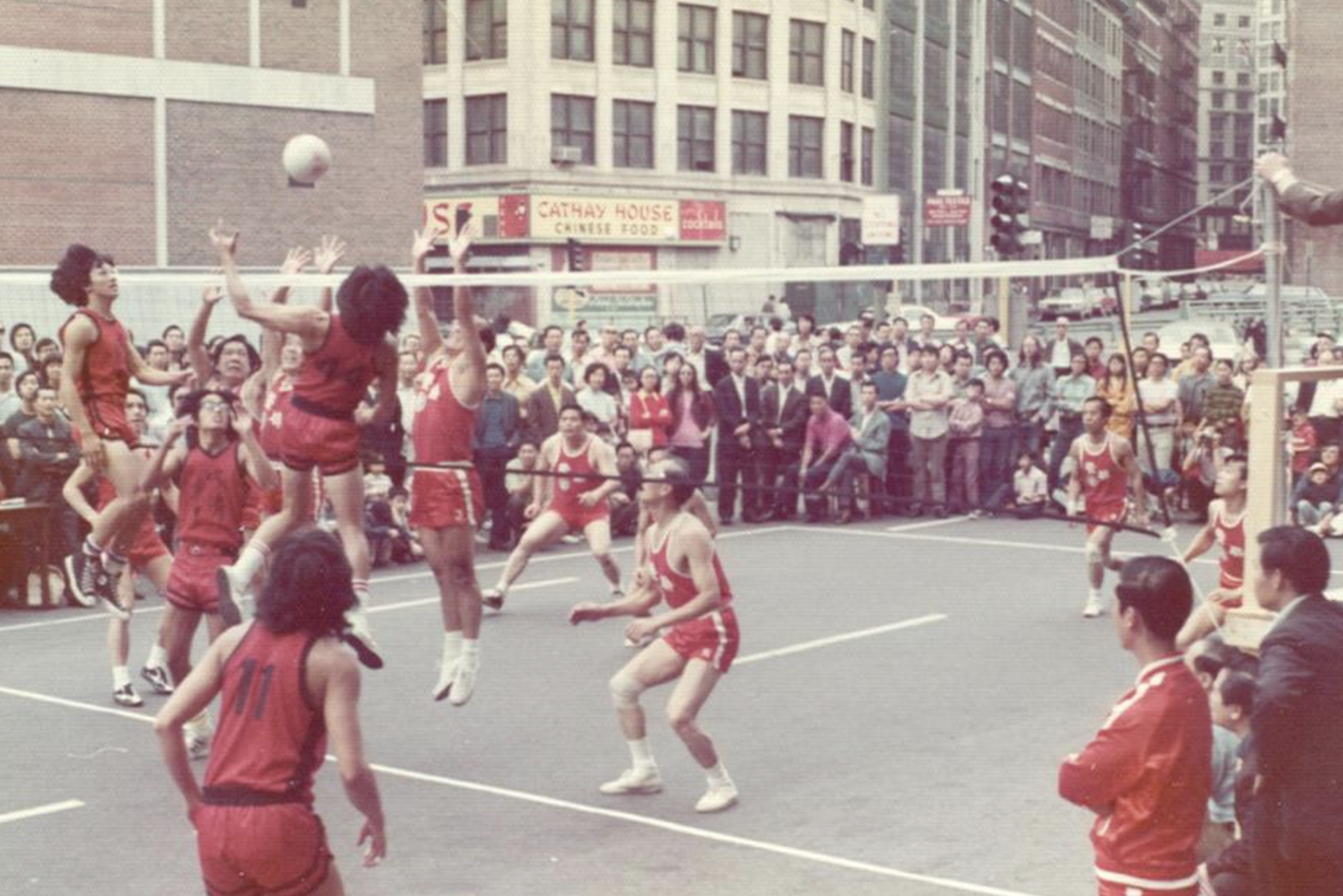A group of women playing volleyball outdoors on a city street, with a crowd of spectators watching behind a barrier, surrounded by tall buildings and storefronts.