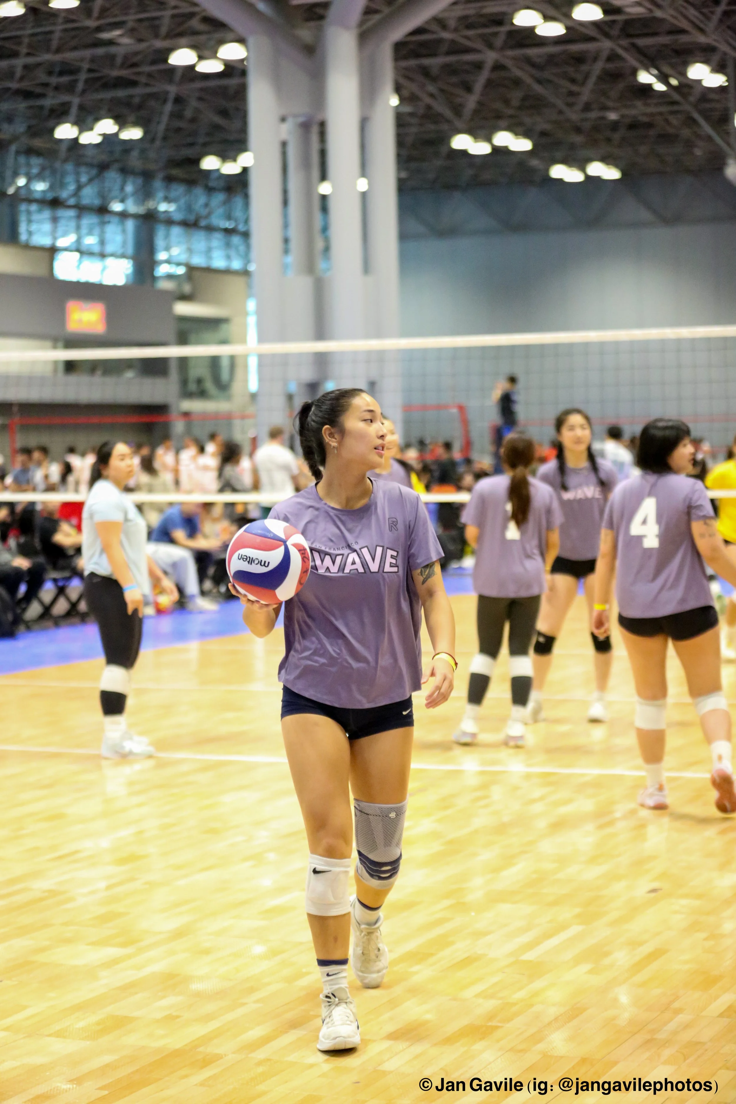 A young woman holding a volleyball standing on an indoor volleyball court, with other players and spectators in the background.