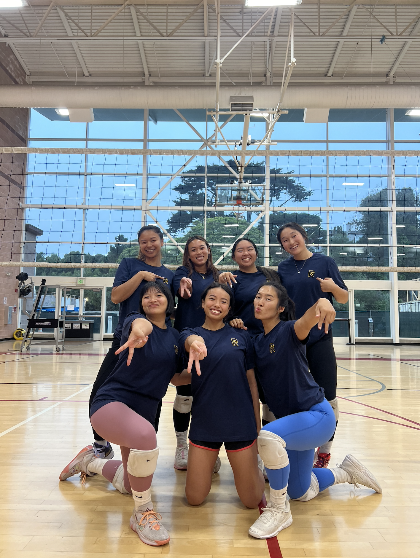 A group of seven young women volleyball players in matching navy blue jerseys and shorts celebrating in a gymnasium with a volleyball net and basketball hoop behind them. Some are kneeling in front, others standing, all smiling and pointing at the camera.