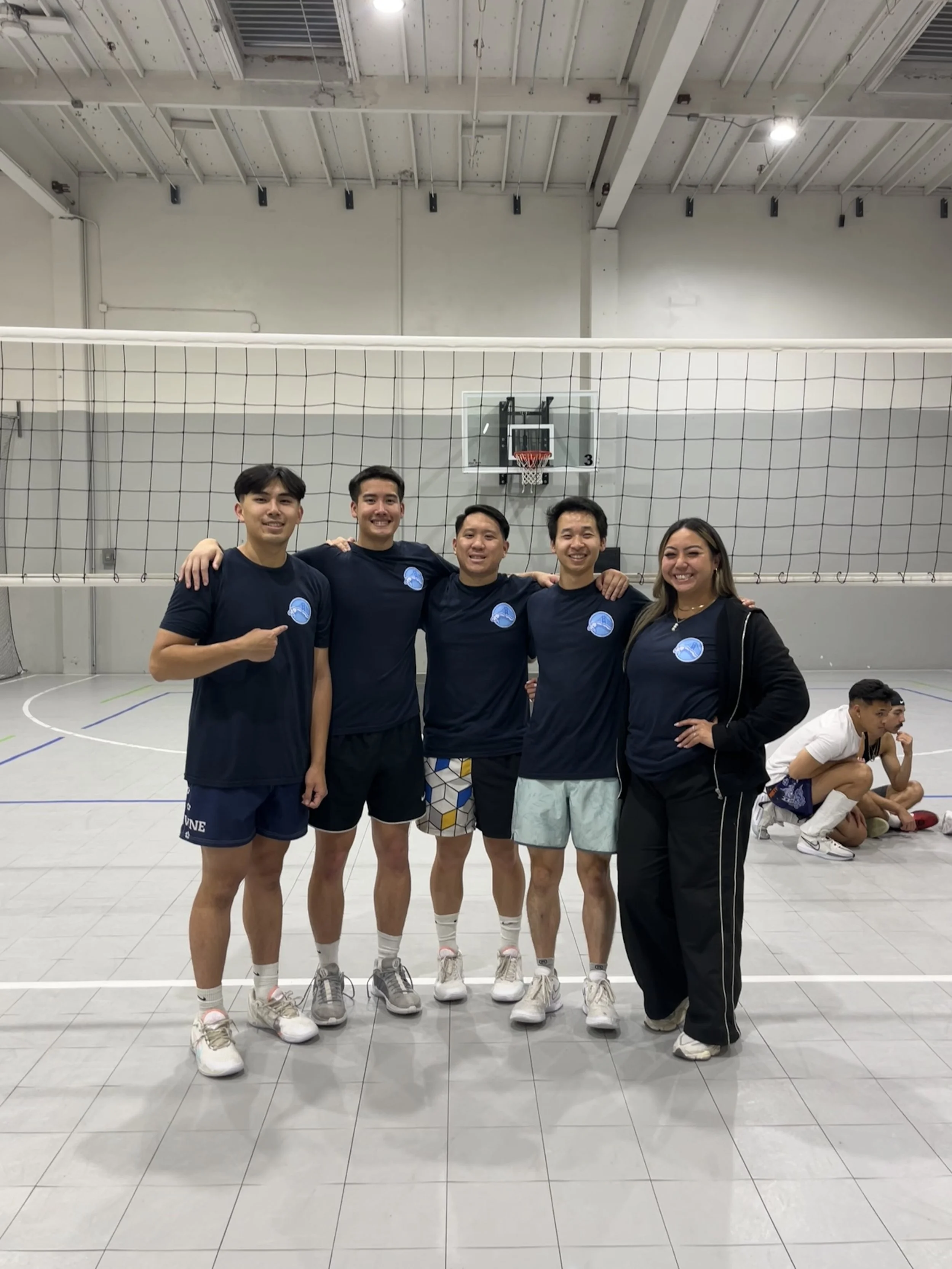 Group of five people standing inside a gymnasium, posing for a photo with their arms around each other, in front of a volleyball net. Four men are wearing dark athletic shirts, shorts, and sneakers, and a woman in a black outfit with a jacket has her