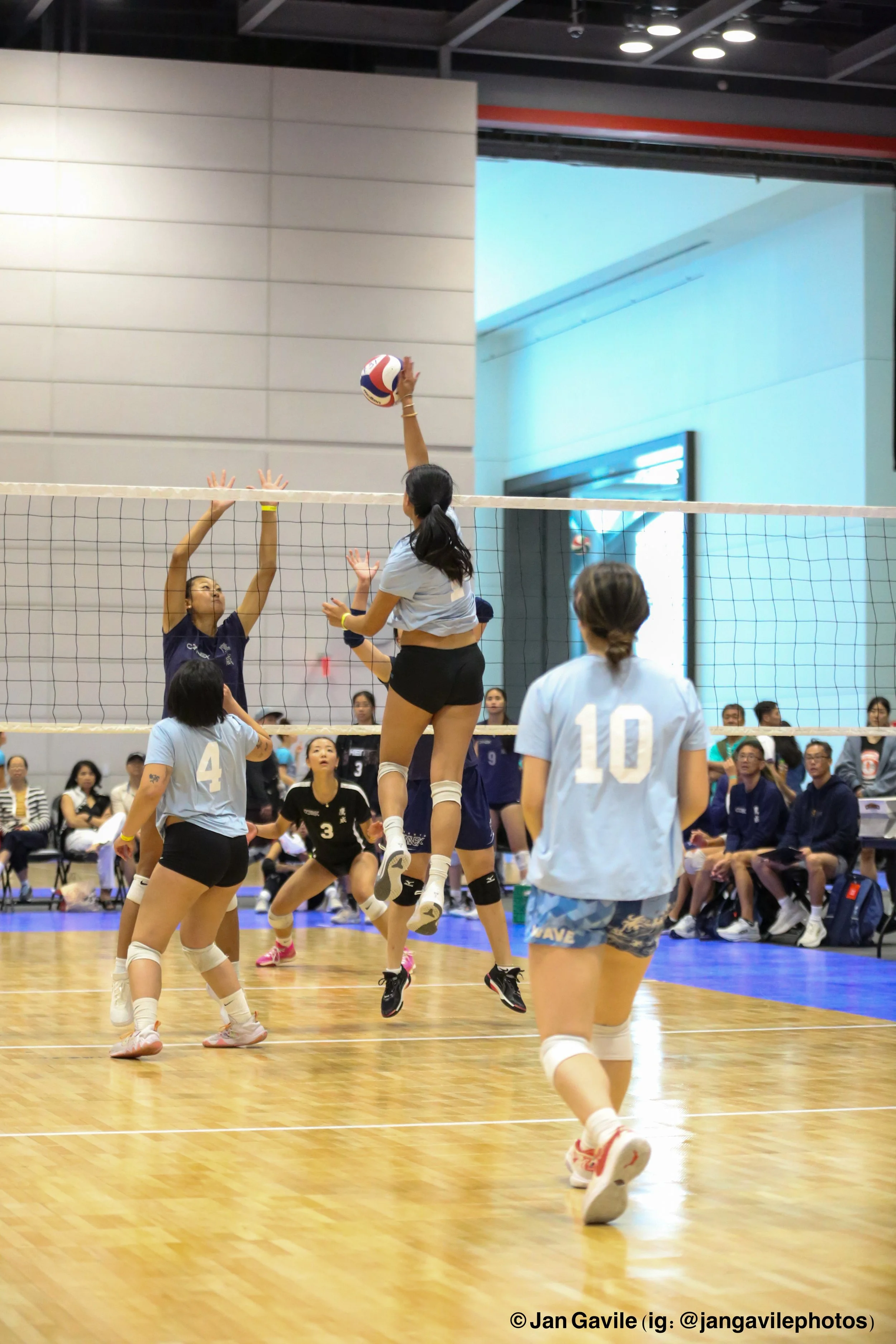 Women playing volleyball in an indoor gym with spectators watching in the background.