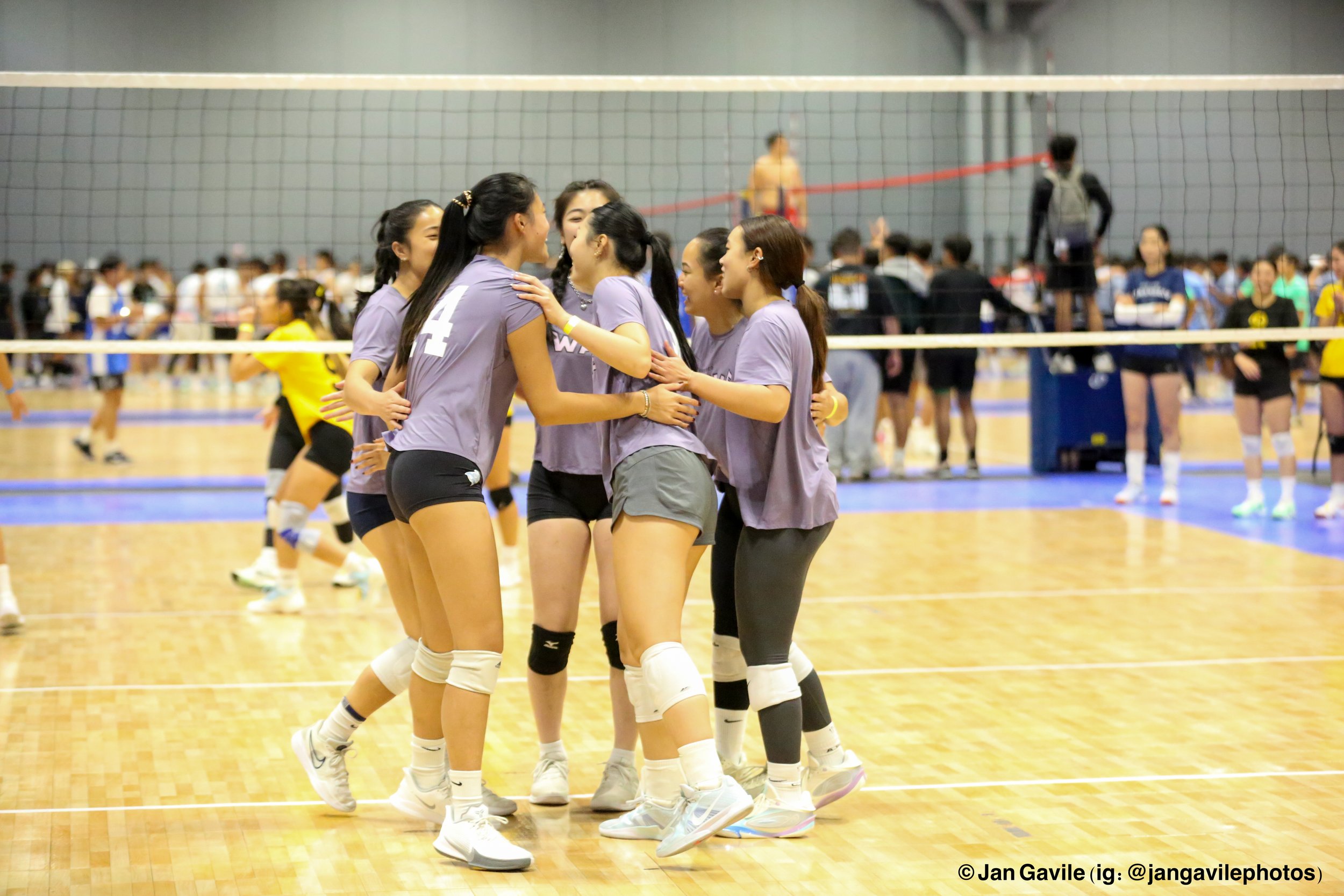 A group of female volleyball players celebrating on an indoor volleyball court. They are wearing purple uniforms and white knee pads, with a volleyball net in the foreground and a crowd in the background.