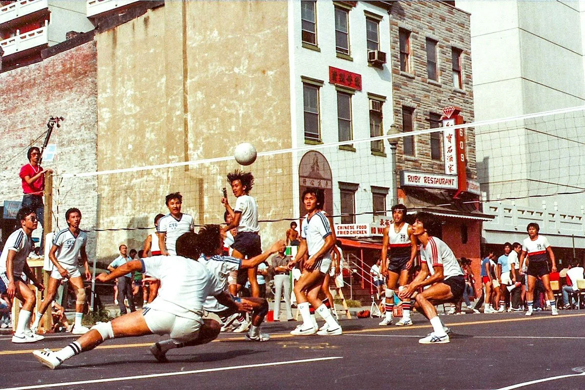 A street volleyball game in an urban setting with players in athletic attire. Some players are jumping, others are ready to receive the ball. The background includes multi-story buildings and a restaurant sign.