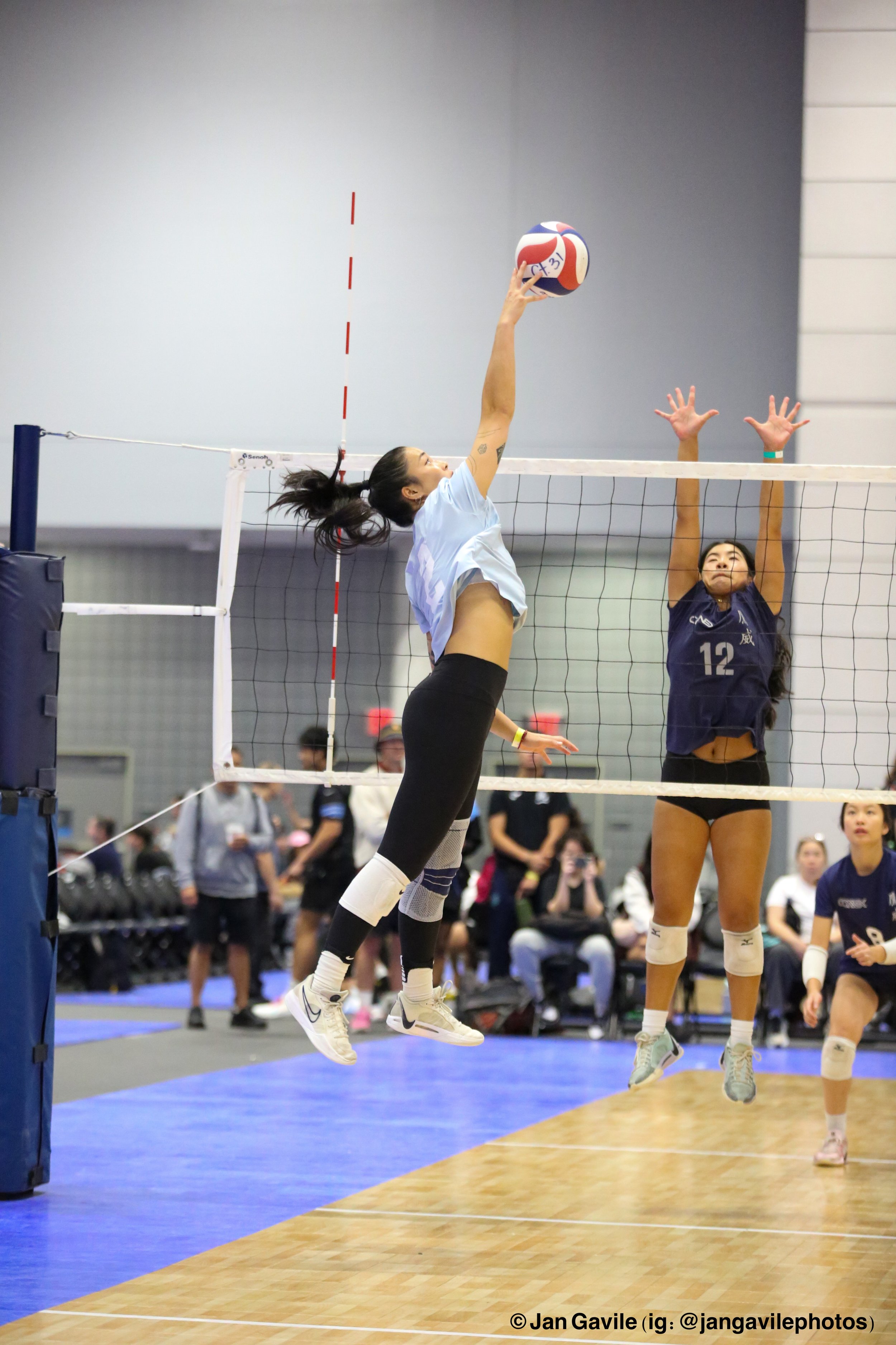 Two female volleyball players in action during a game; one is jumping to hit the ball, while the other is prepared to block at the net; spectators watch in the background.