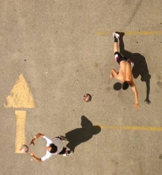 Two boys playing volleyball on an outdoor concrete court, one shirtless, the other in a white shirt, with shadows cast on the ground.