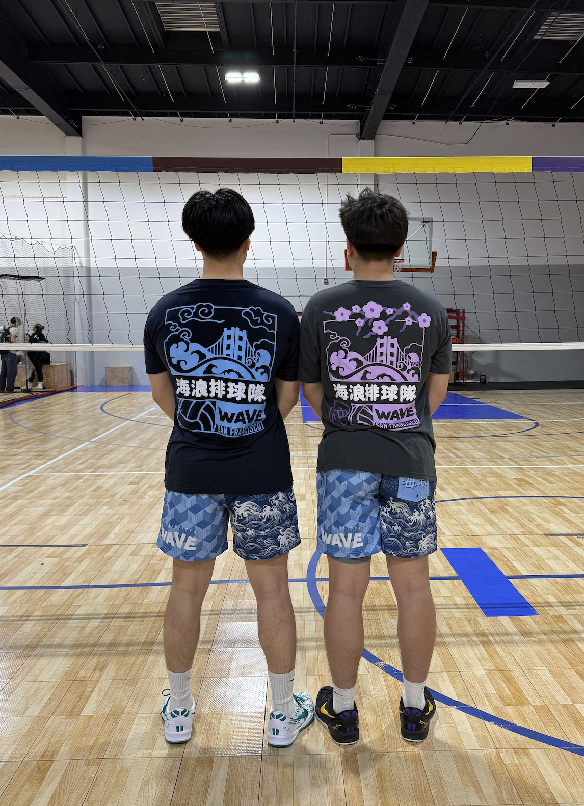 Two young men standing on a gymnasium volleyball court, facing away from the camera. They are wearing matching t-shirts with graphic designs and the word 'WAVE' and shorts with wave patterns. The volleyball net and a basketball hoop are visible in th