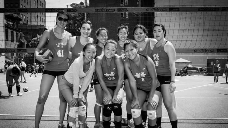 Girls' volleyball team posing on an outdoor court with a net, with some players kneeling and others standing, holding a volleyball, during daytime.