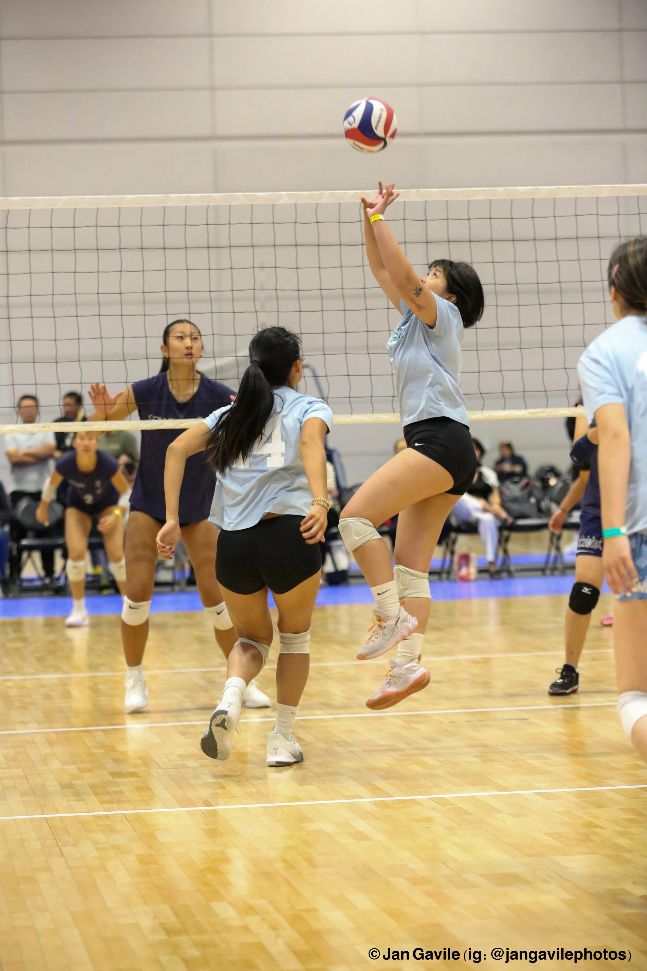A group of young women playing volleyball indoors. One woman is jumping up to hit the ball over the net, while others are preparing to follow the play. The background shows a gymnasium with spectators seated and watching the game.