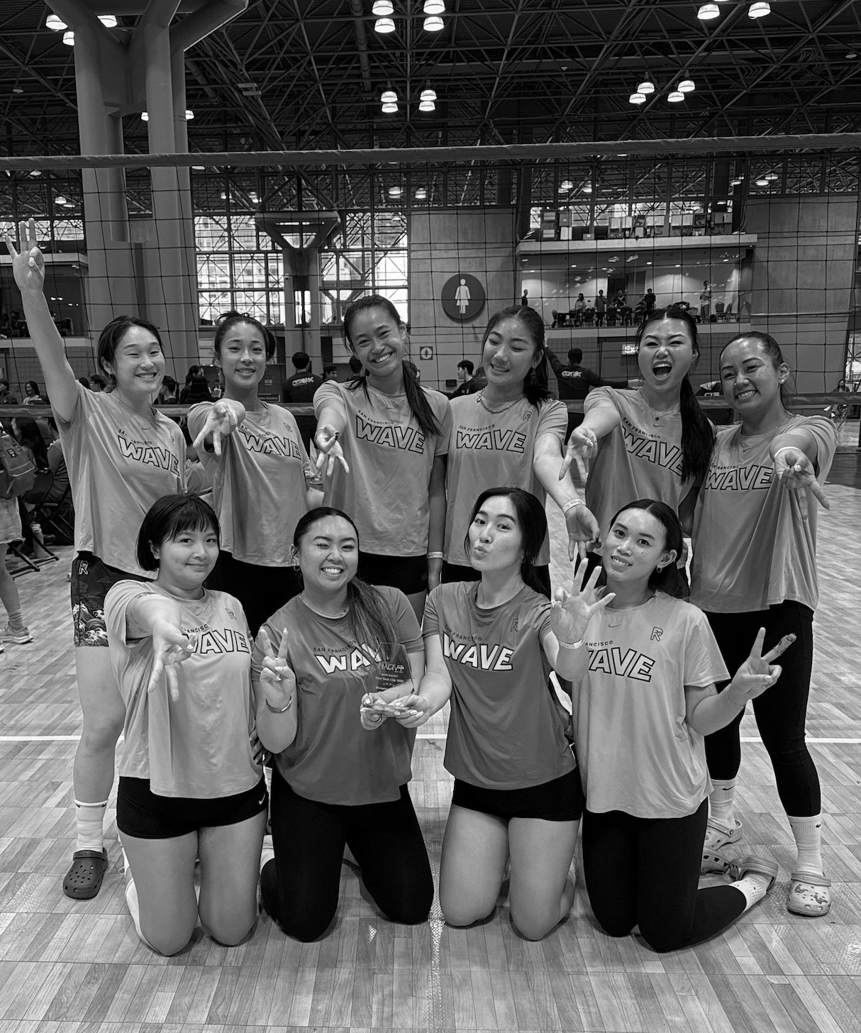 A group of nine female volleyball players in sports uniforms posing together inside an indoor sports arena, smiling and making various hand gestures, with a volleyball net and bleachers in the background.